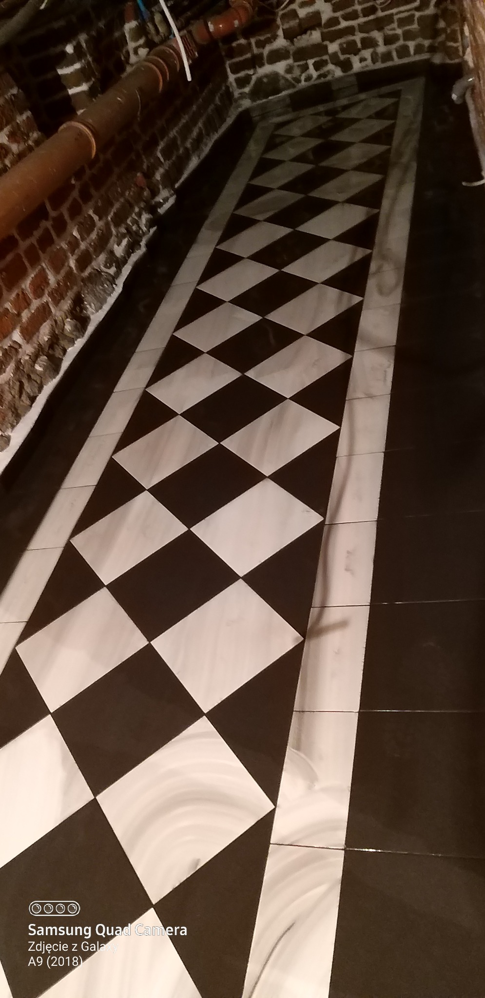 Ukończona podłoga w łazience z czarno-białą szachownicą z płytek, widoczne rury przy ścianie z cegły. Nietypowe wykończenie.