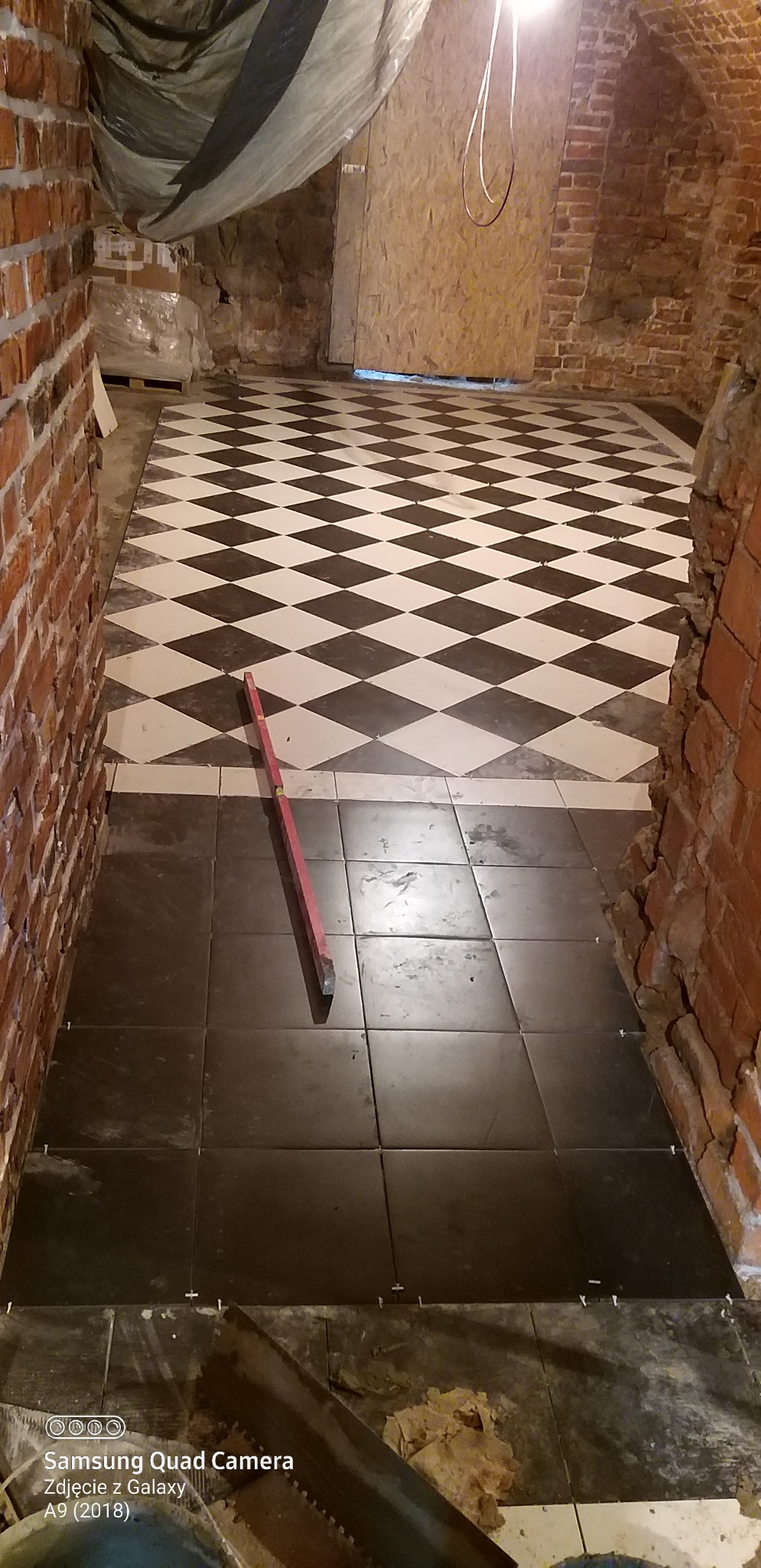 Układanie ciemnych płytek podłogowych w piwnicy z fragmentem podłogi w szachownicę; poziomica na płytkach, widoczne krzyżyki dystansowe i ceglane ściany.