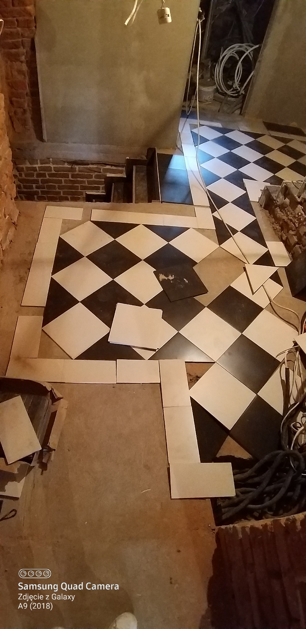 Układanie czarno-białej podłogi z płytek w romby, widoczne niedokończone krawędzie i przygotowane płytki. W tle fragment ceglanej ściany i instalacje.