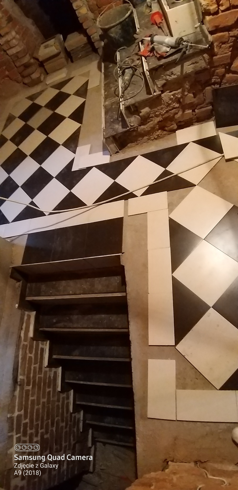 Układanie czarno-białej szachownicy z płytek na podłodze i schodach w trakcie remontu. Widoczne narzędzia i materiały budowlane. Detal wykończenia wnętrza.