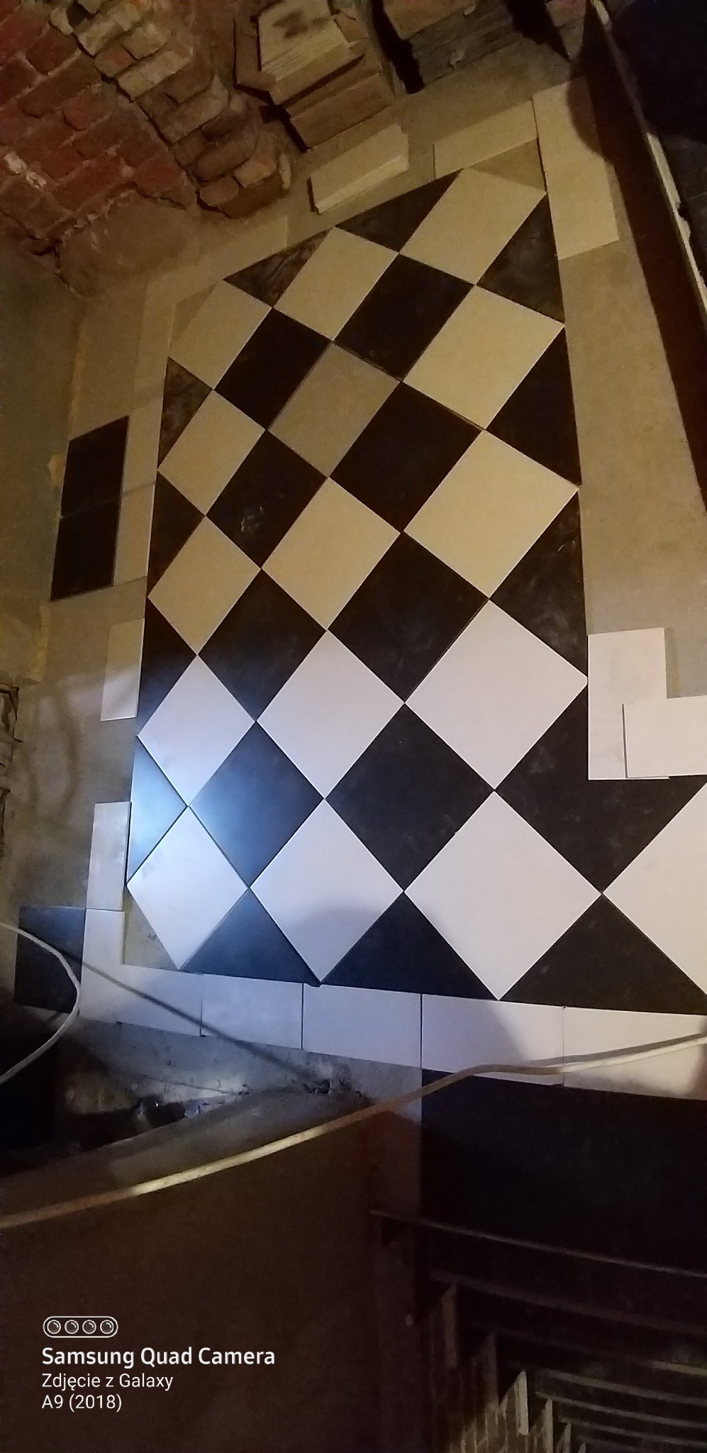 Układanie czarno-białej podłogi w karo. Widoczny fragment ceglanego sufitu i niedokończone krawędzie. Użyto płytek o kwadratowym kształcie.