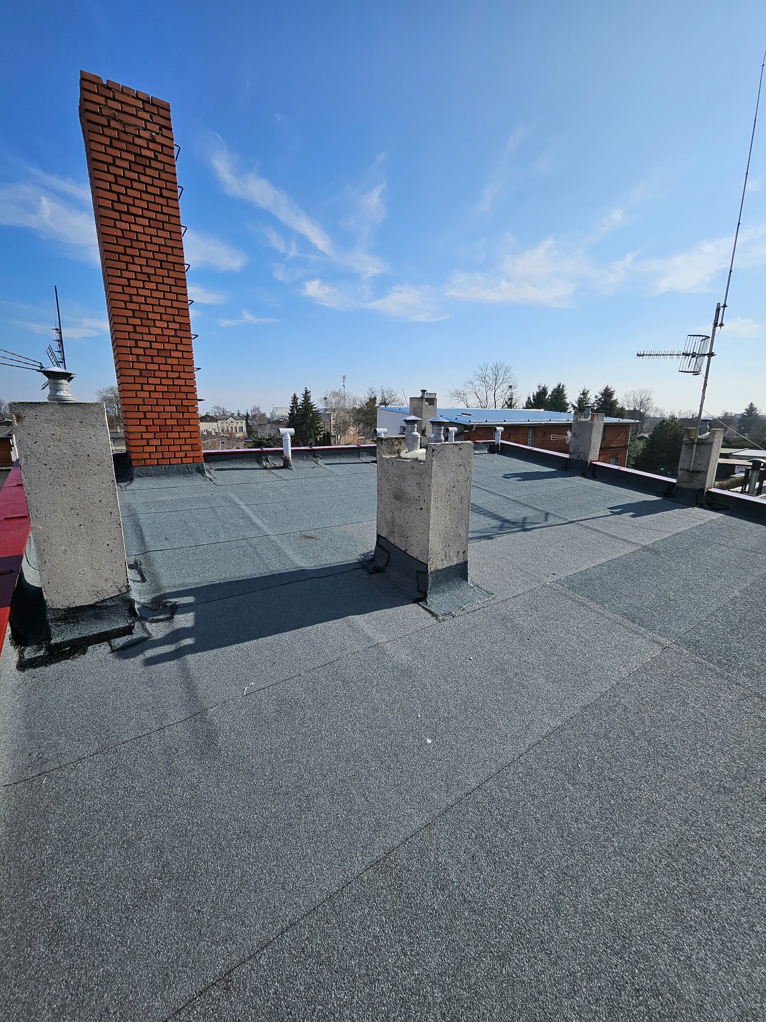 Szary dach pokryty papą termozgrzewalną z kominem z cegły, betonowymi elementami i antenami na tle błękitnego nieba. Widok z góry na płaską powierzchnię dachu.