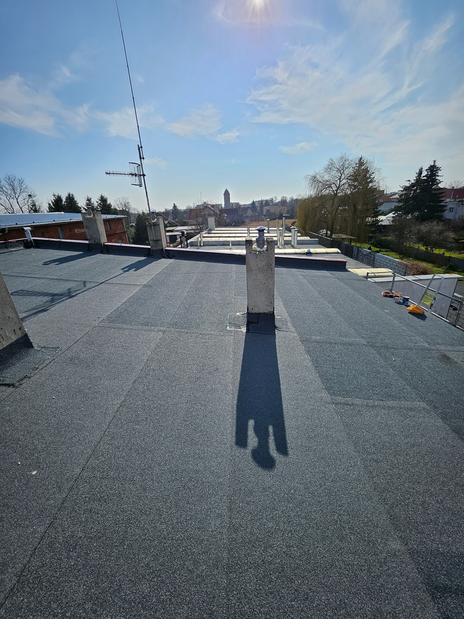 Widok z góry na płaski dach pokryty nową, ciemną papą termozgrzewalną w słoneczny dzień. W tle kominy, antena i zabudowania.