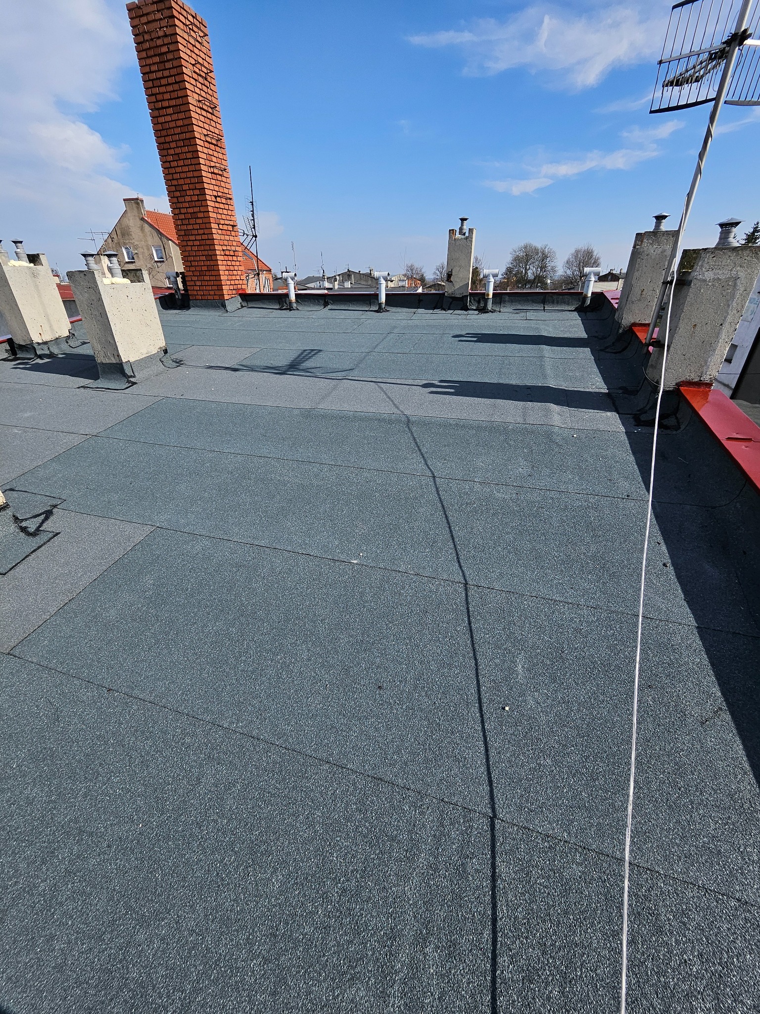 Szary dach pokryty papą termozgrzewalną w słoneczny dzień. Widoczne kominy, anteny i fragmenty budynków w tle. Detal pokrycia dachowego.