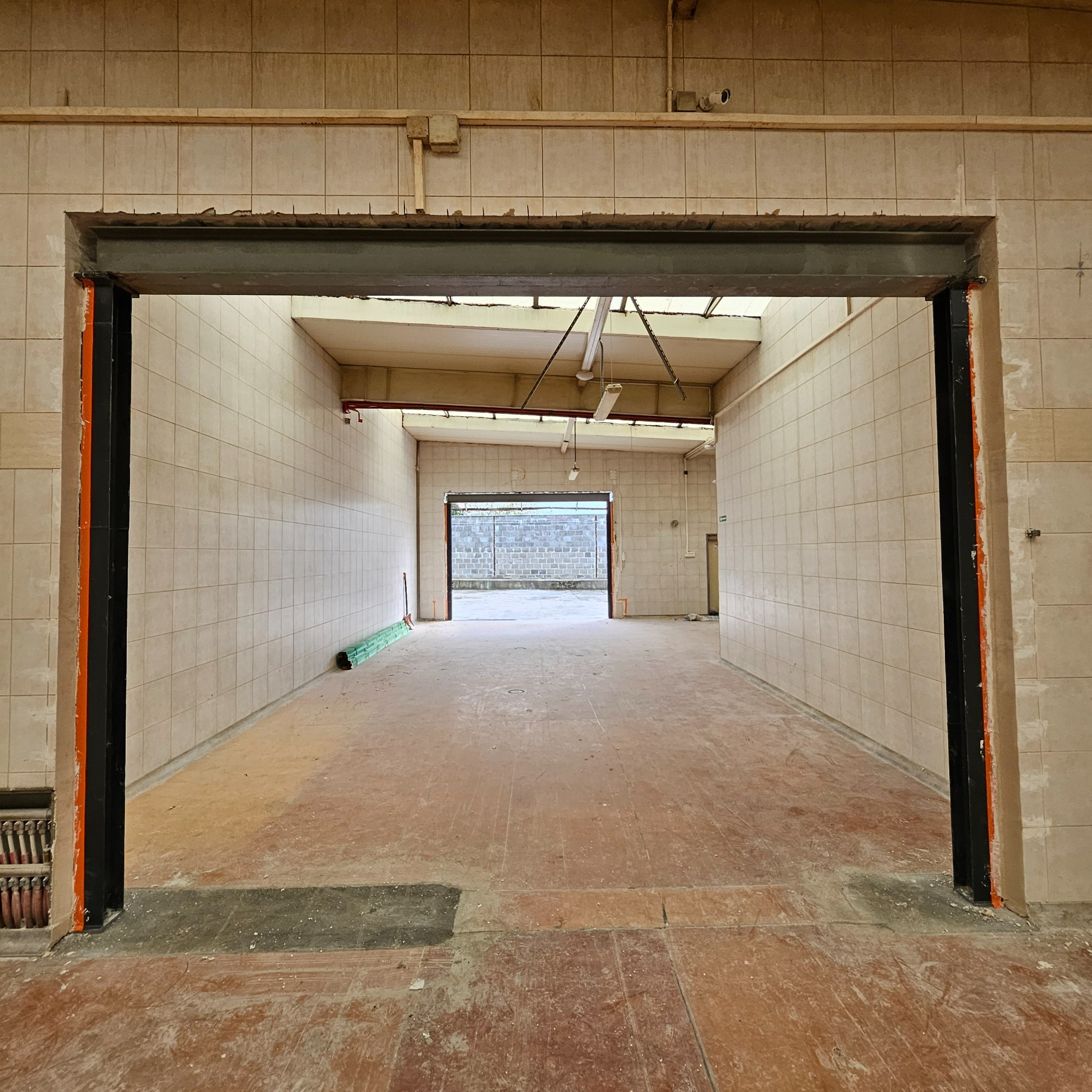Wnętrze w trakcie remontu: beżowe płytki na ścianach, surowe podłoże, stalowa rama drzwiowa z pomarańczowymi elementami. Widok na otwartą przestrzeń z bramą w tle.