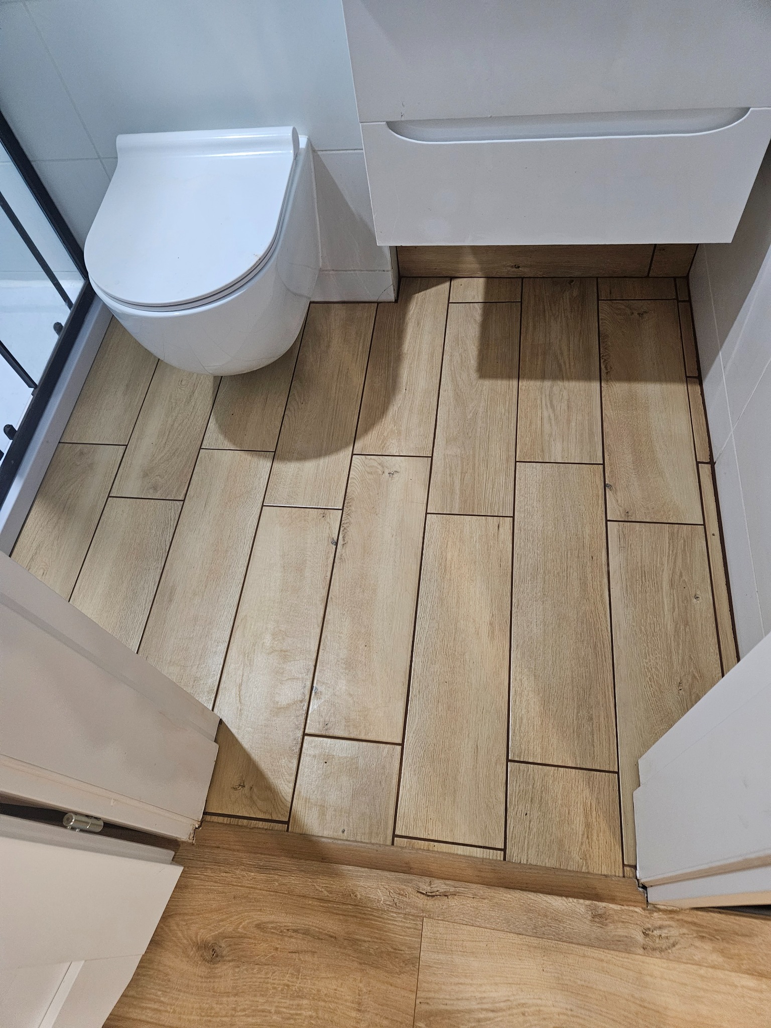 Wykończona łazienka z toaletą, szafką umywalkową i kabiną prysznicową. Na podłodze ułożone płytki imitujące drewno. Widok z góry, z otwartych drzwi.