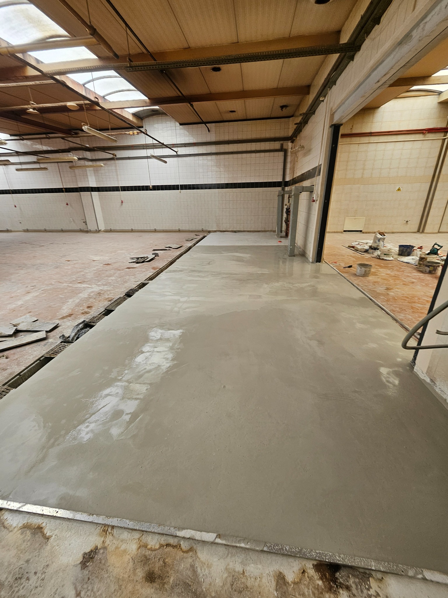 Wylewka samopoziomująca w jasnym, przestronnym wnętrzu z widocznymi śladami wcześniejszych prac. Częściowo wykończona podłoga, surowe ściany i sufit z oknami dachowymi.