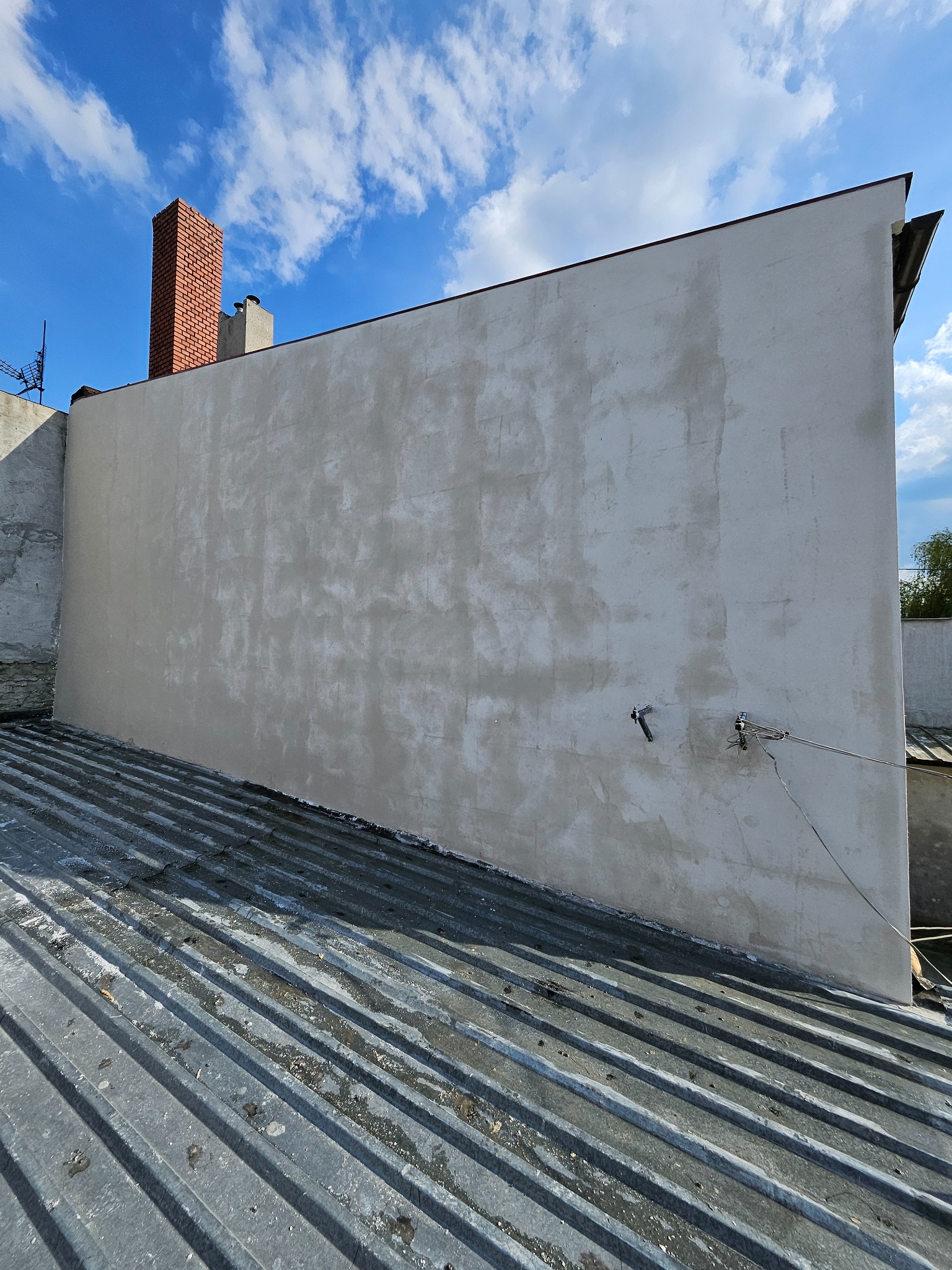 Świeżo otynkowana ściana budynku z widocznym kominem z czerwonej cegły na tle błękitnego nieba z chmurami. Widok z dachu z blachy trapezowej.