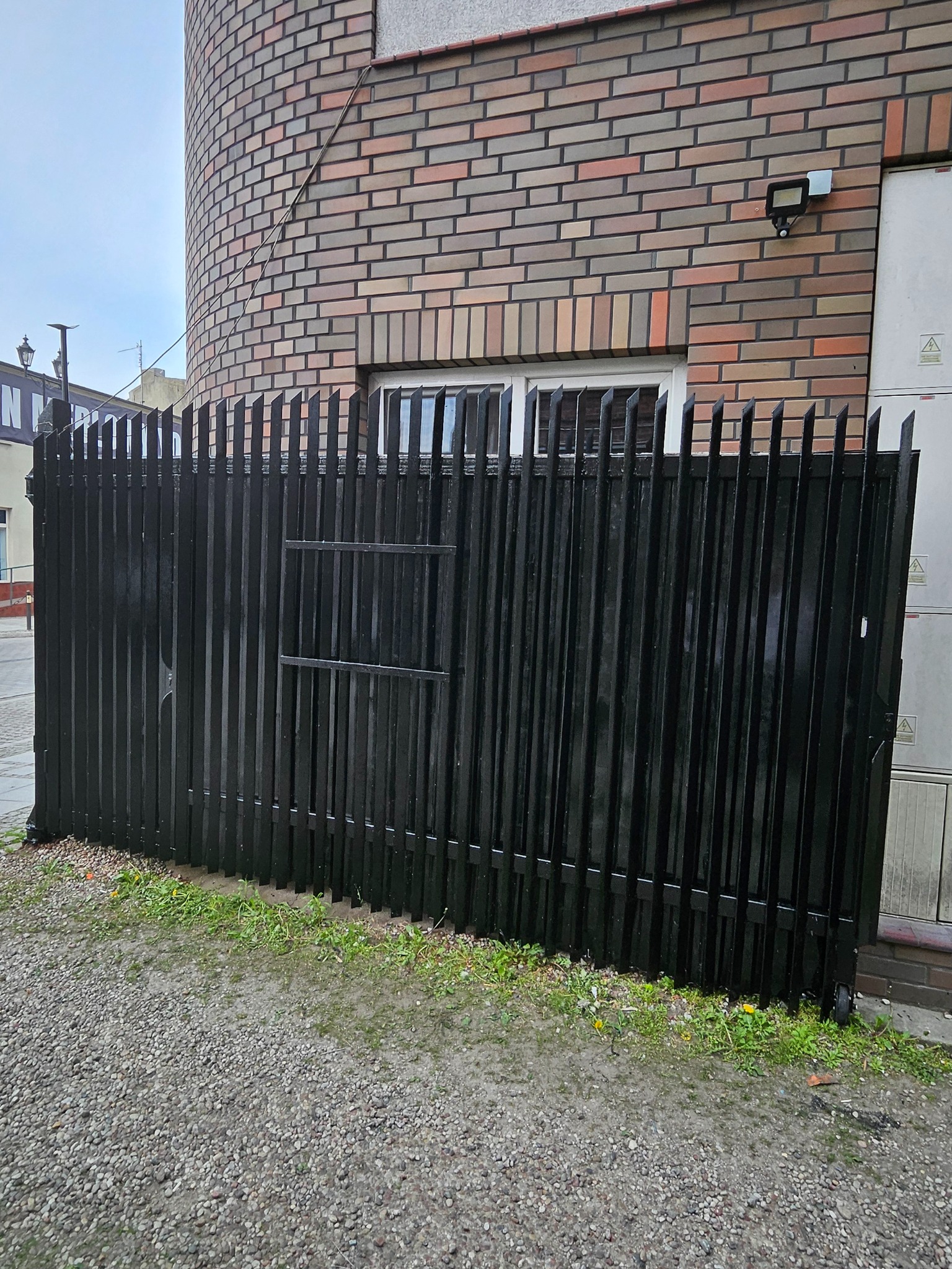Czarny, metalowy płot z bramką na tle ceglanej ściany budynku z oknem. Widoczne kamienie i trawa u podstawy ogrodzenia.