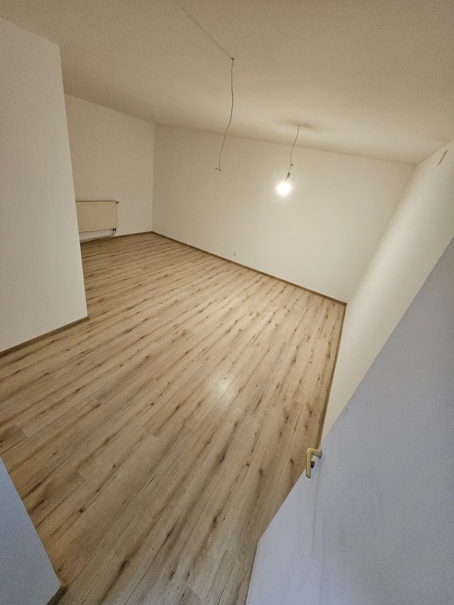 Jasne panele podłogowe w pustym pokoju z białymi ścianami i grzejnikiem. Widoczne kable zwisające z sufitu. Perspektywa z narożnika pokoju.
