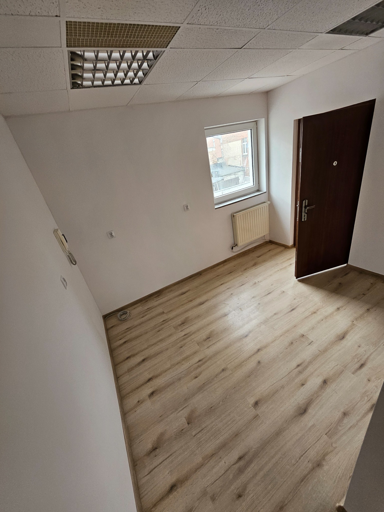 Puste, jasne wnętrze biurowe z panelami podłogowymi, grzejnikiem pod oknem i brązowymi drzwiami. Widoczna instalacja elektryczna i kratka wentylacyjna na suficie.