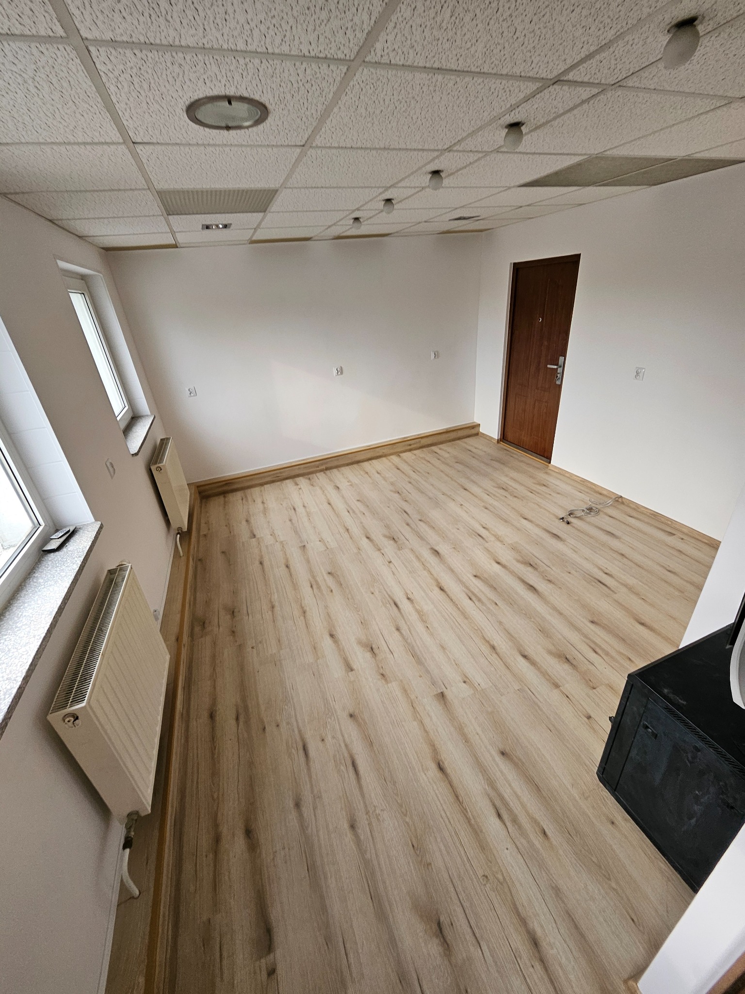 Jasne panele podłogowe w pustym pokoju z białymi ścianami i grzejnikami. Widoczny fragment sufitu podwieszanego i brązowe drzwi. Neutralne oświetlenie.