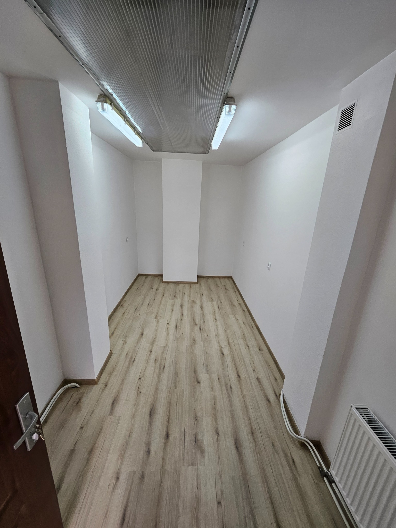 Wnętrze odnowionego pomieszczenia biurowego z drewnianą podłogą, białymi ścianami i sufitami z oświetleniem rastrowym. Widok z perspektywy drzwi wejściowych.