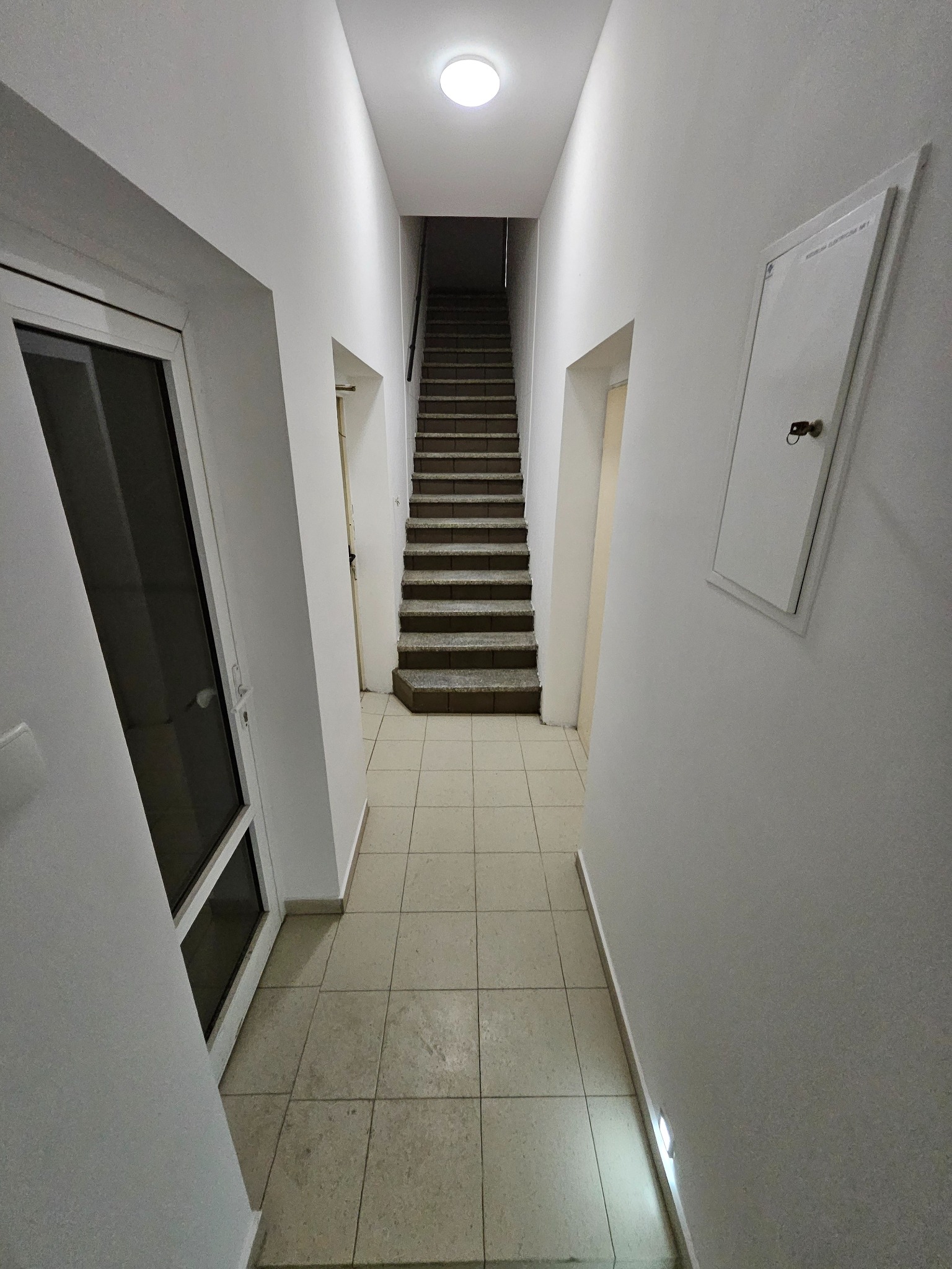 Wnętrze klatki schodowej z białymi ścianami i beżową podłogą z płytek. Widoczne schody, drzwi i skrzynka elektryczna na ścianie. Oświetlenie sufitowe.