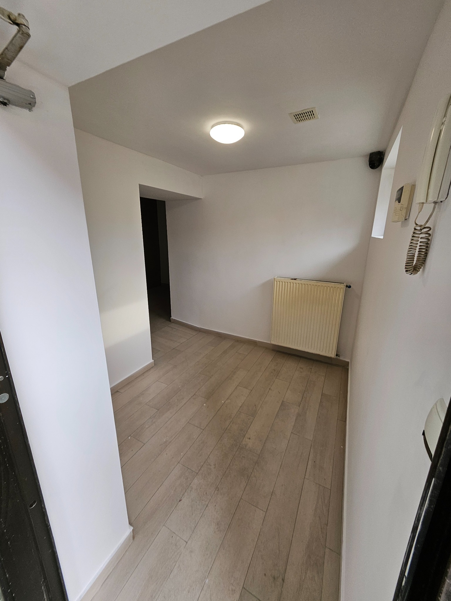 Jasne, wykończone wnętrze korytarza z drewnopodobną podłogą, białymi ścianami i grzejnikiem. Widoczny fragment drzwi wejściowych i domofon. Nowoczesne oświetlenie sufitowe.