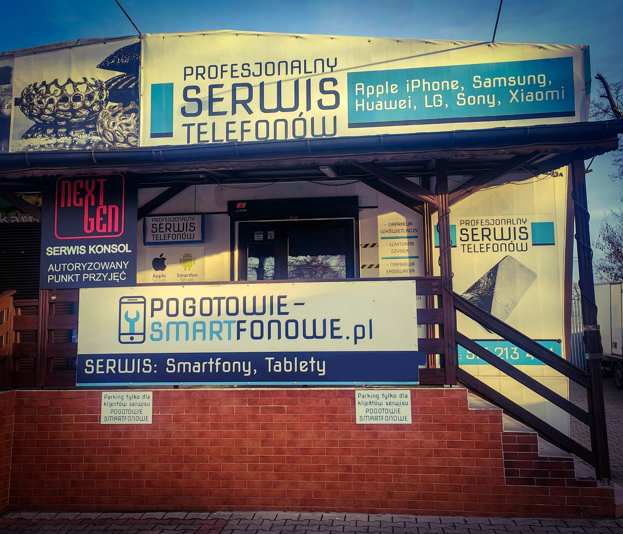 Fasada punktu serwisowego telefonów z szyldami informacyjnymi o naprawie smartfonów różnych marek. Widoczne logo i adres strony internetowej.
