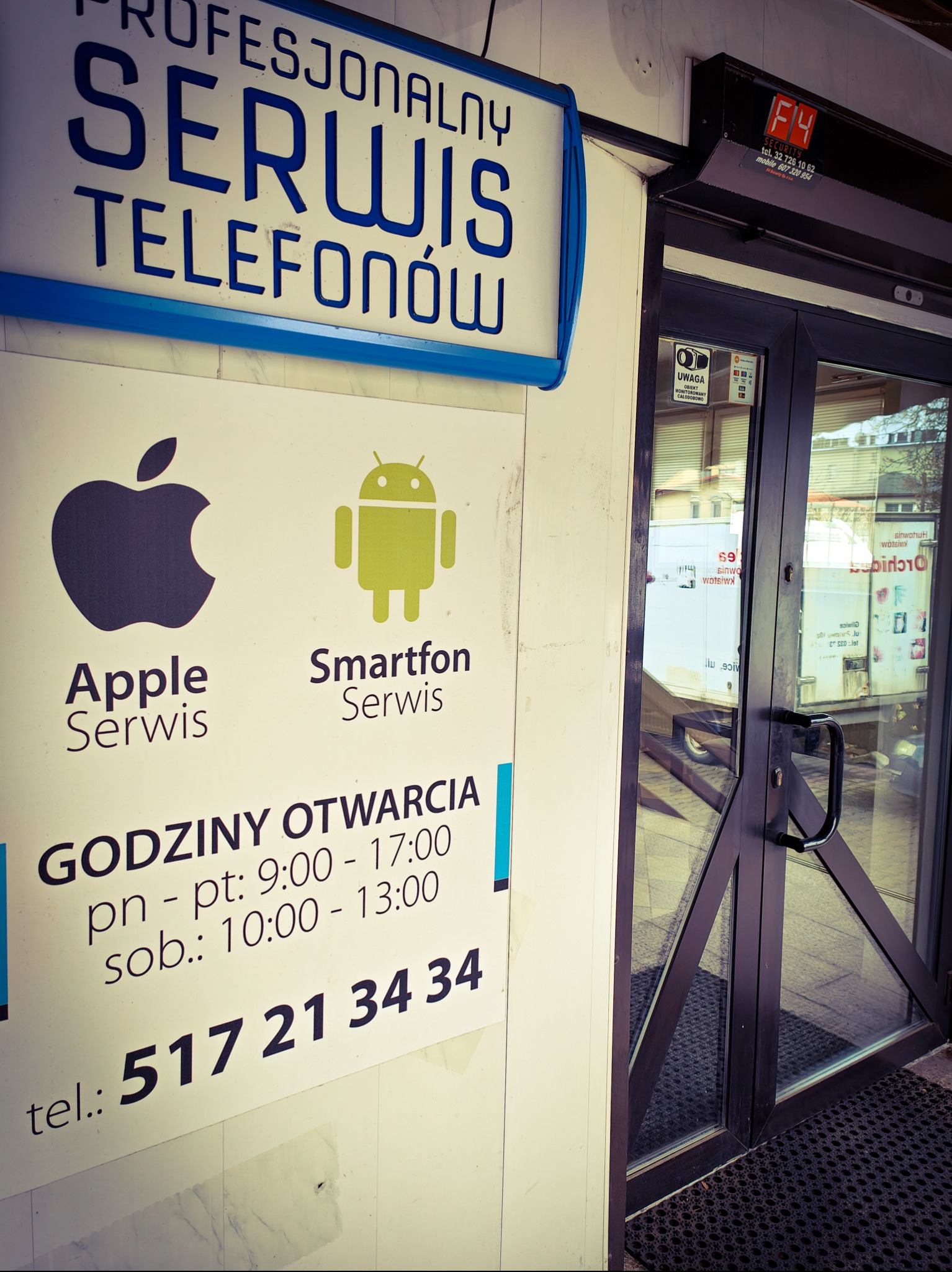 Wejście do serwisu telefonów z logo Apple i Android. Tabliczka z godzinami otwarcia i numerem telefonu. Nad wejściem szyld 'Profesjonalny Serwis Telefonów'.