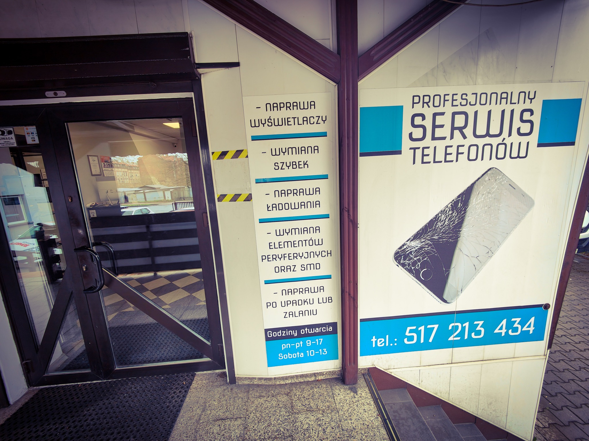 Wejście do serwisu telefonów z plakatem informującym o naprawie wyświetlaczy, wymianie szybek i ładowaniu. Widoczny numer telefonu kontaktowego i godziny otwarcia.