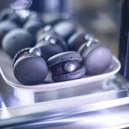 Ciemne, artystyczne makaroniki z błyszczącymi srebrnymi akcentami na białym talerzu w chłodnej witrynie, prezentowane w cukierni.