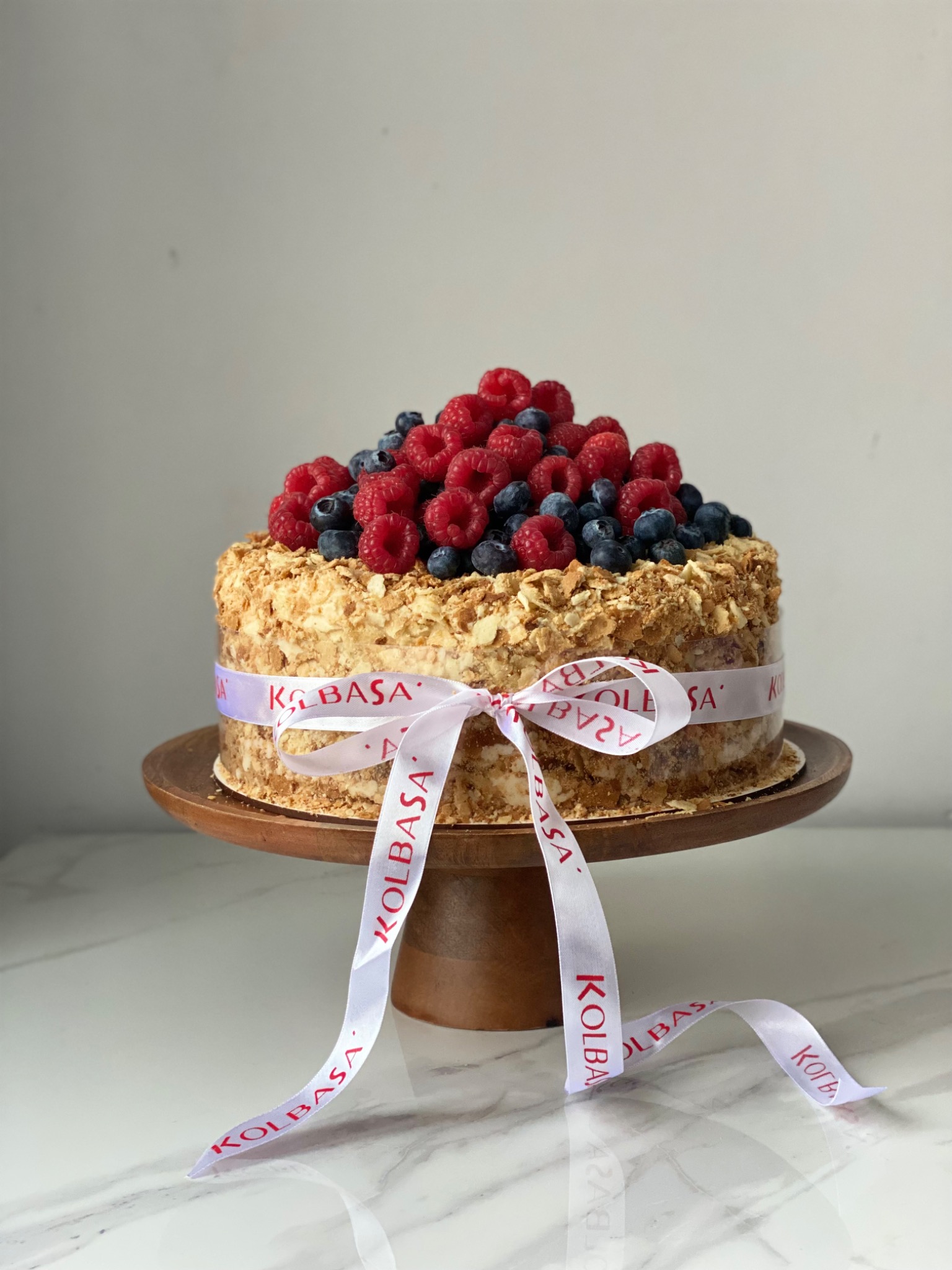 Tort Napoleon na drewnianej paterze, udekorowany malinami i borówkami, przewiązany białą wstążką z czerwonym napisem 'KOLBASA'. Kremowe wnętrze widoczne przez przezroczystą obudowę.