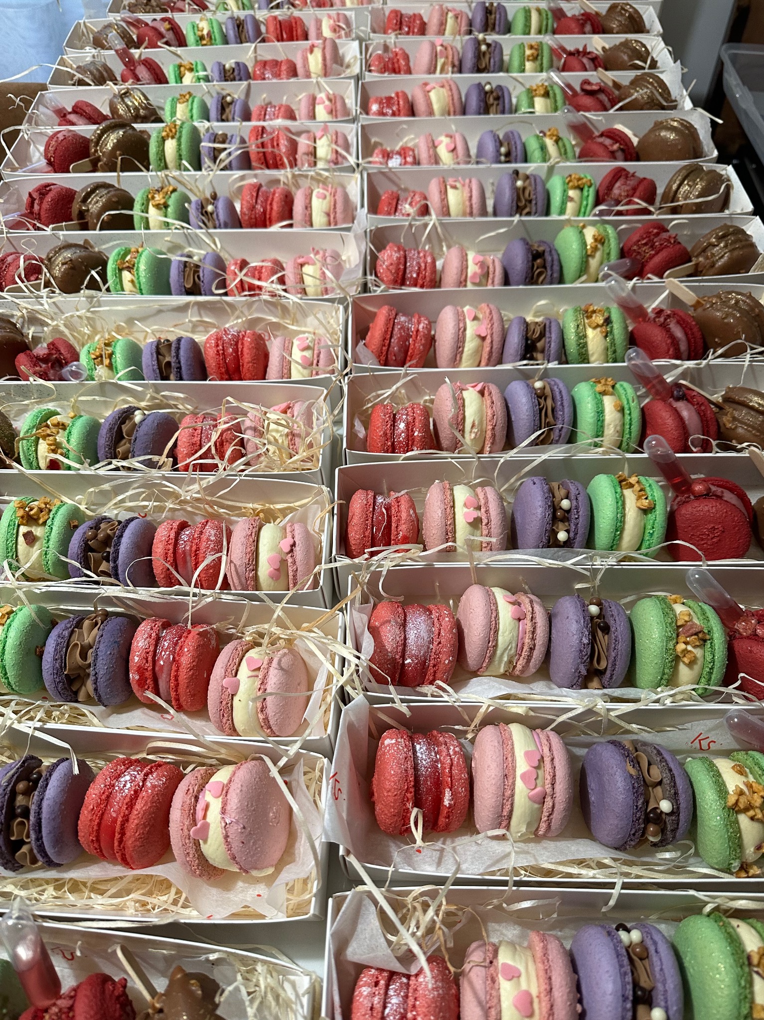 Rzędy pudełek z kolorowymi makaronikami o różnych smakach i dekoracjach, ułożone jeden na drugim. Widok z góry ukazuje bogactwo oferty.