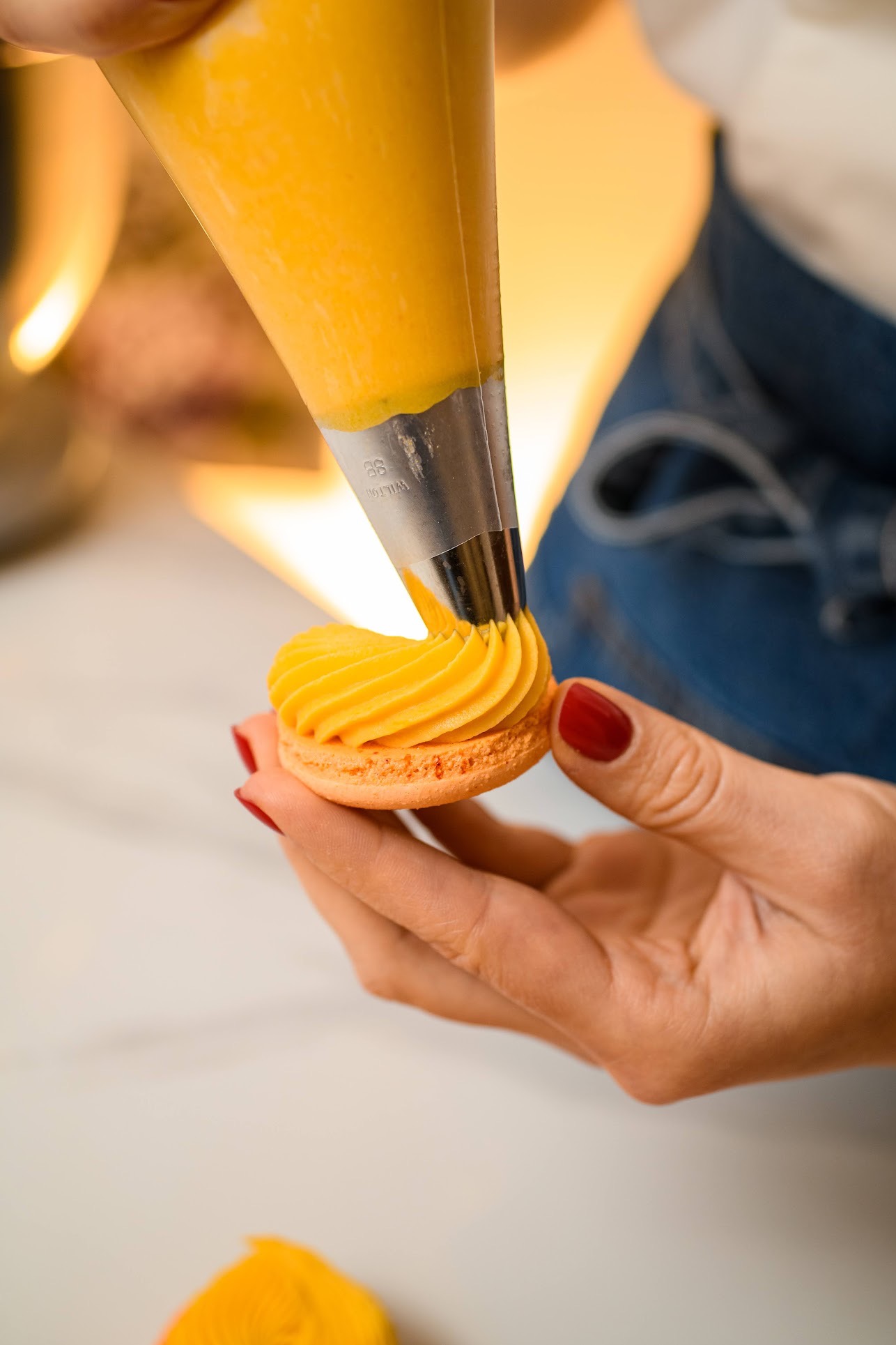 Dekorowanie pomarańczowego makaronika żółtym kremem z rękawa cukierniczego, widoczne dłonie z czerwonym manicure, delikatne oświetlenie.