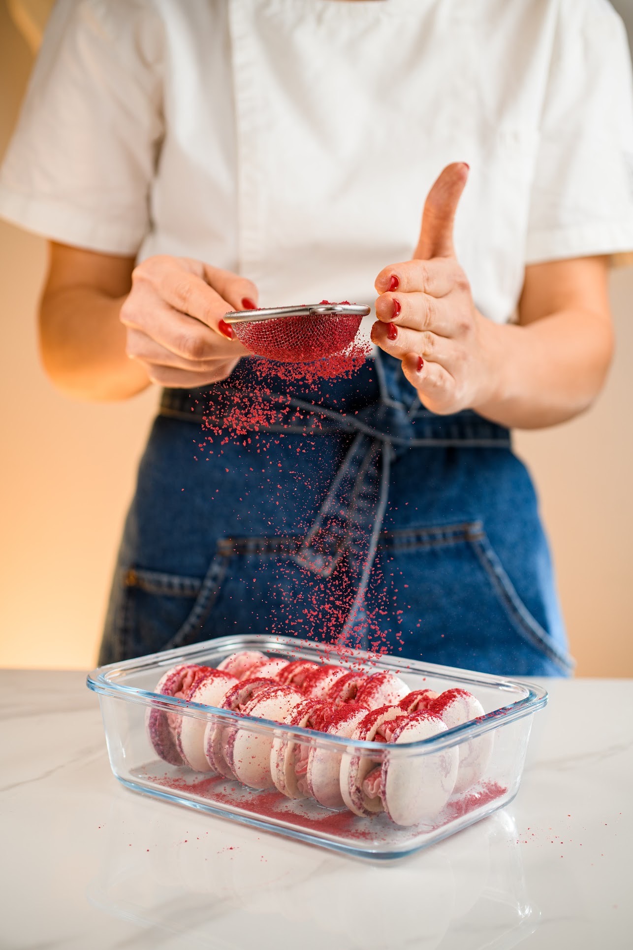 Kucharz posypuje różowym pudrem makaroniki w szklanym naczyniu, tworząc artystyczny efekt wizualny. Widoczne dłonie z czerwonym manicure, biały fartuch i dżinsowy kombinezon.