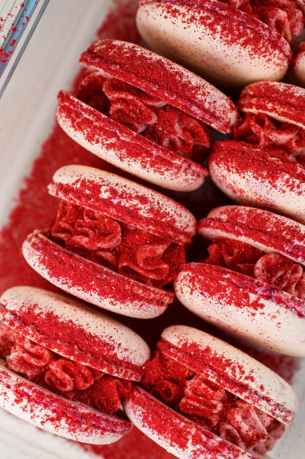 Makaroniki w pudełku, posypane czerwonym proszkiem. Kremowe nadzienie, zbliżenie na detale tekstury i koloru. Delikatne, pastelowe barwy.