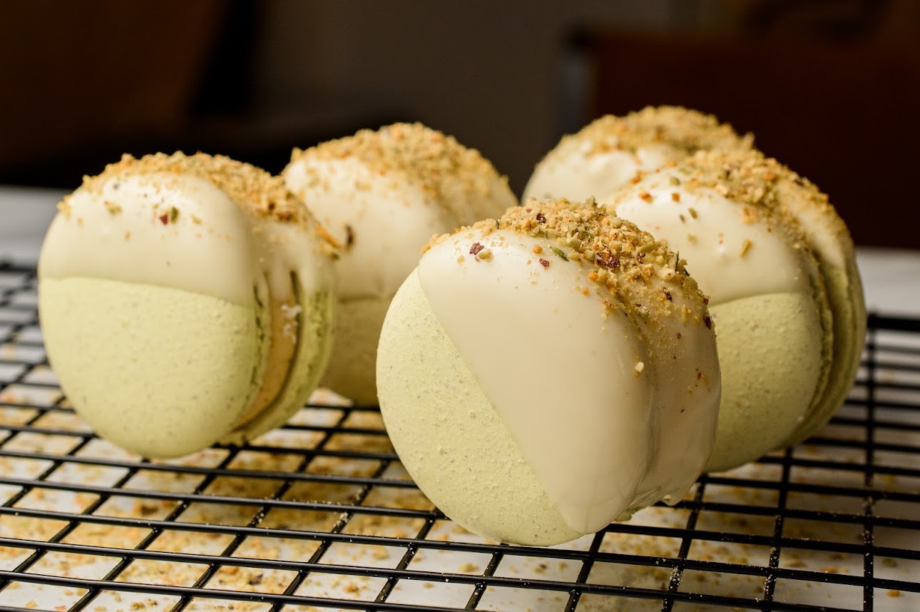 Makaroniki pistacjowe z białą polewą i posypką na metalowej kratce. Delikatne światło podkreśla teksturę i kolory.