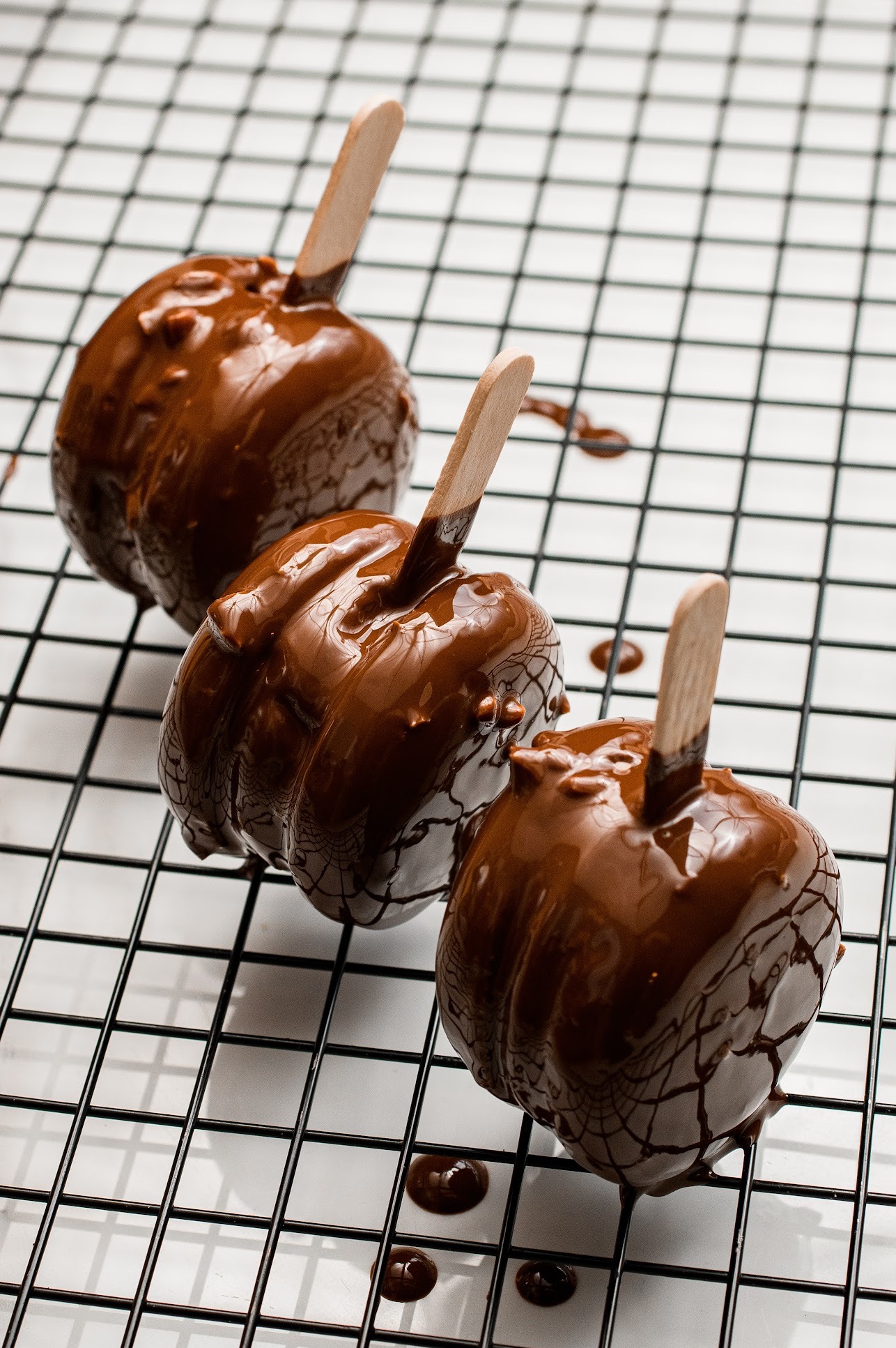 Trzy jabłka oblane czekoladą na patyku, ułożone rzędem na metalowej kratce. Widoczne krople czekolady. Błyszcząca polewa, równomierne pokrycie.