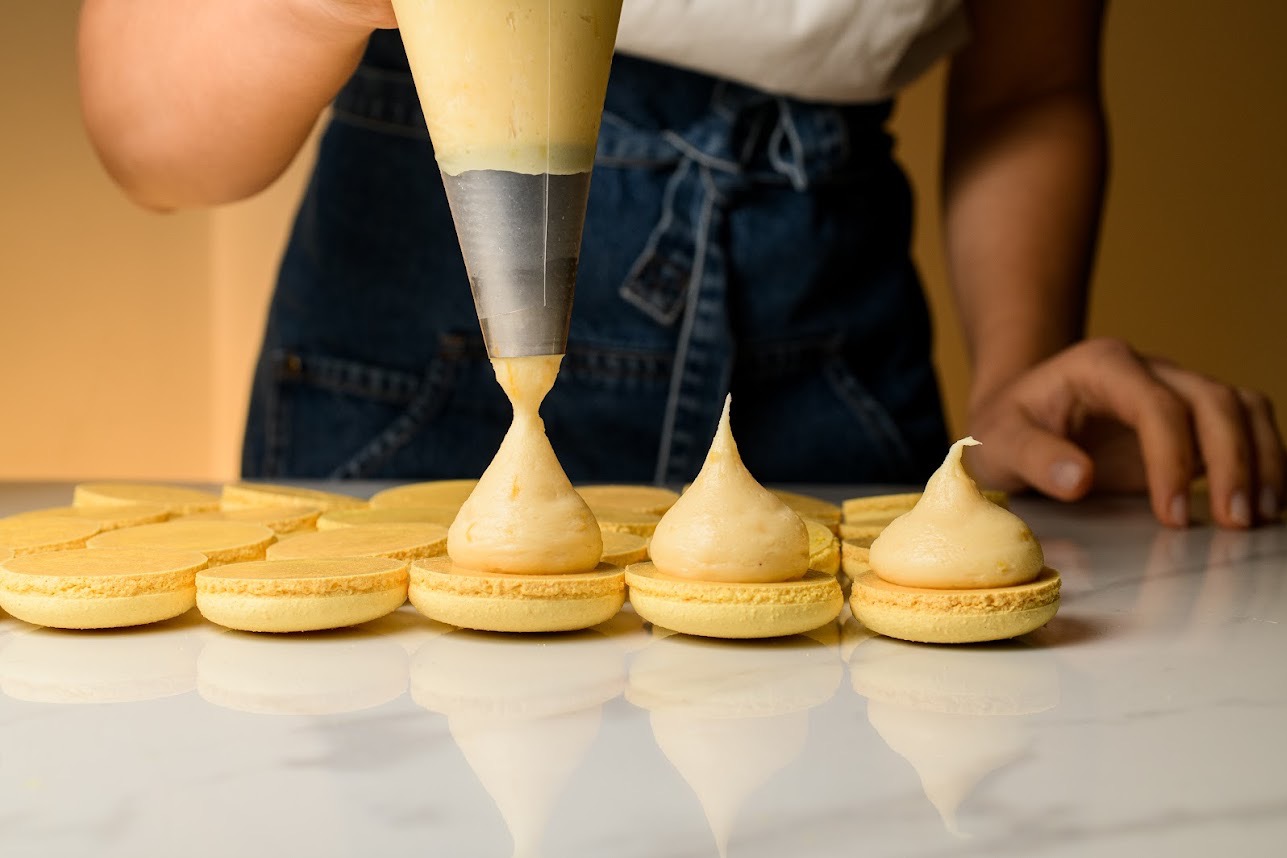 Nakładanie kremu z rękawa cukierniczego na żółte makaroniki. W tle widoczna osoba w dżinsowej bluzie. Blat odbija światło.