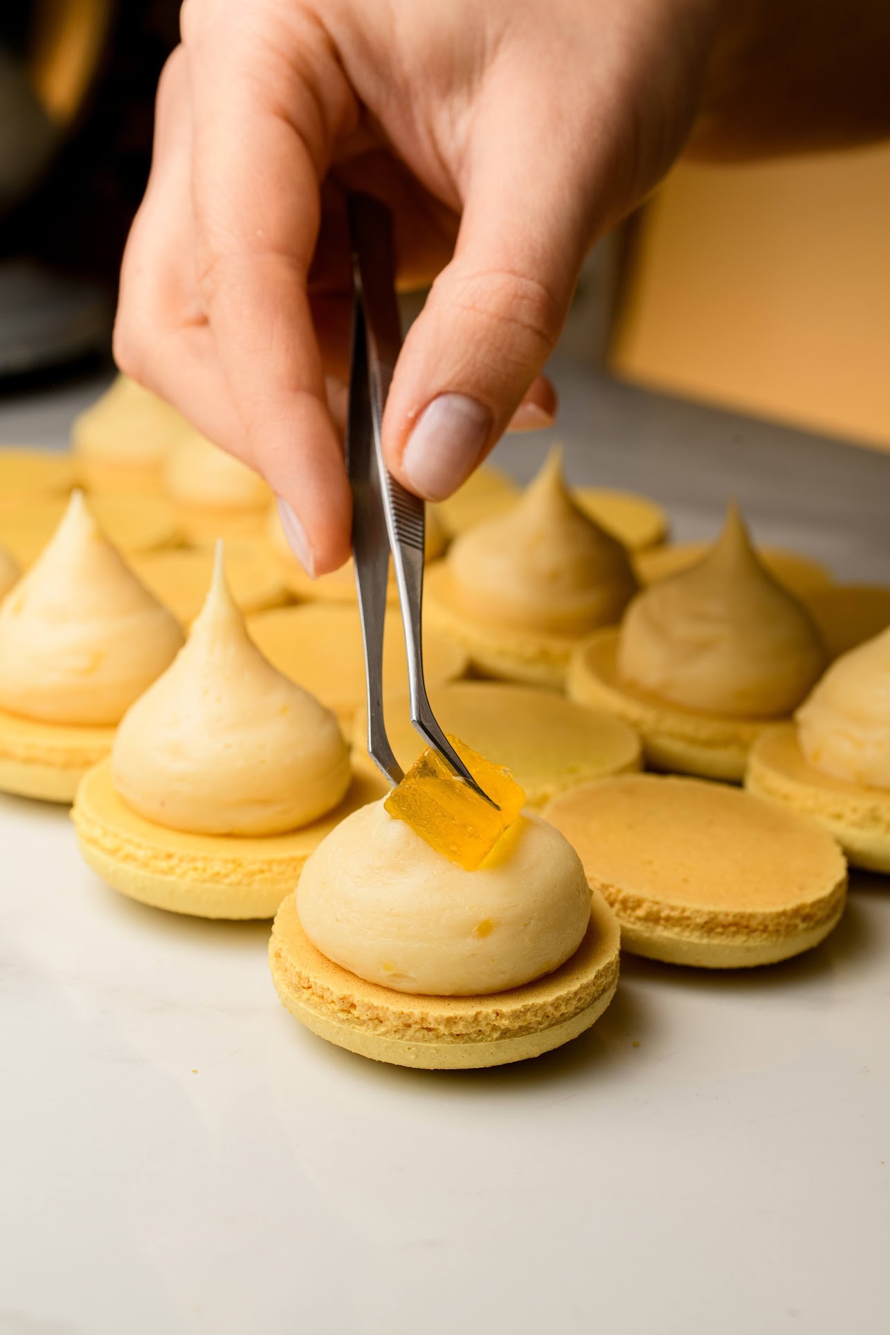 Precyzyjne dekorowanie makaronika z kremem i galaretką przy użyciu pęsety. Pastelowa kolorystyka, bliski kadr, skupienie na detalu wykończenia.