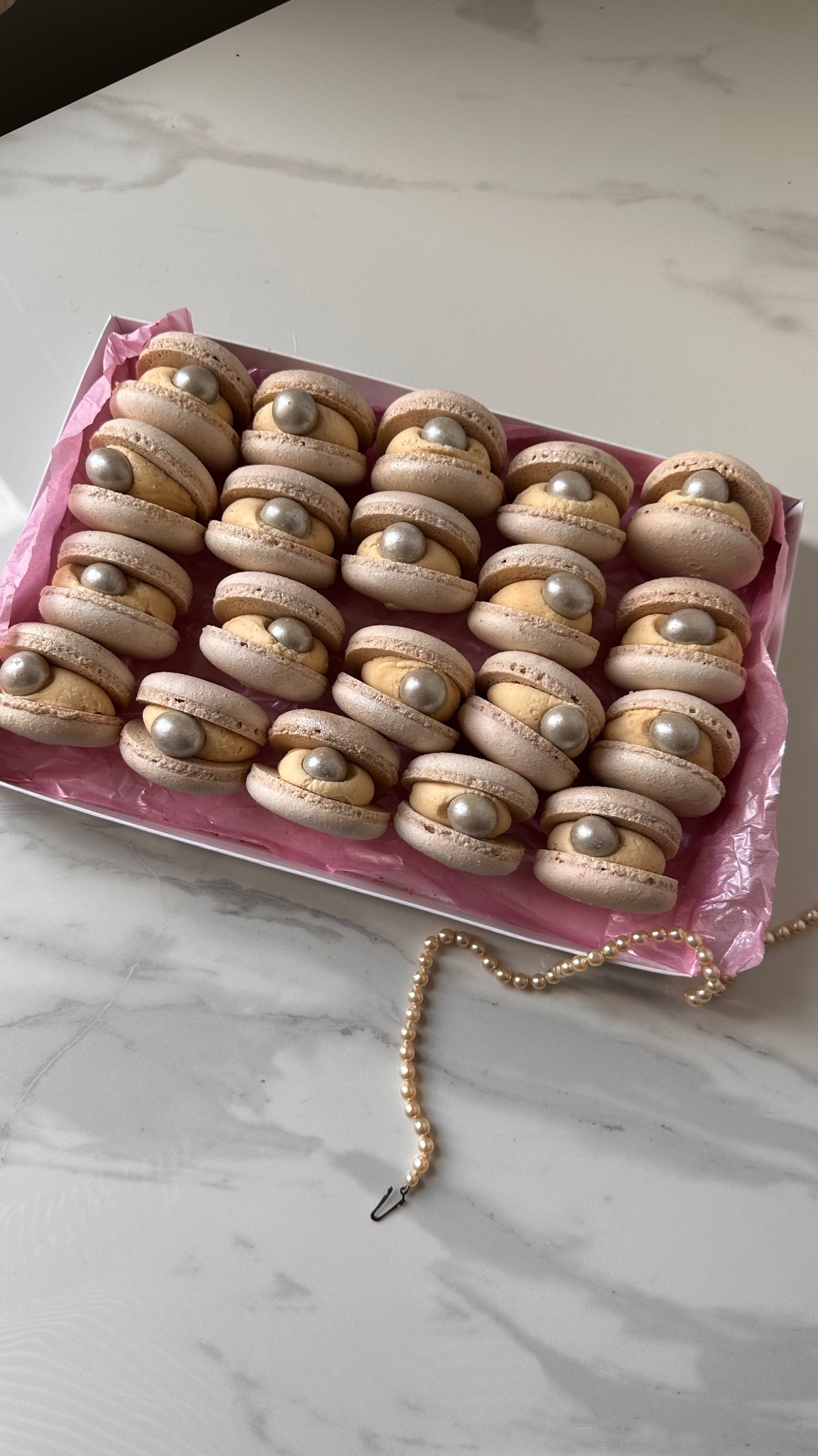Unikatowe makaroniki w kształcie muszelek z perłami w srebrnym kolorze, ułożone w różowym pudełku, obok leży sznur pereł na marmurowym tle.
