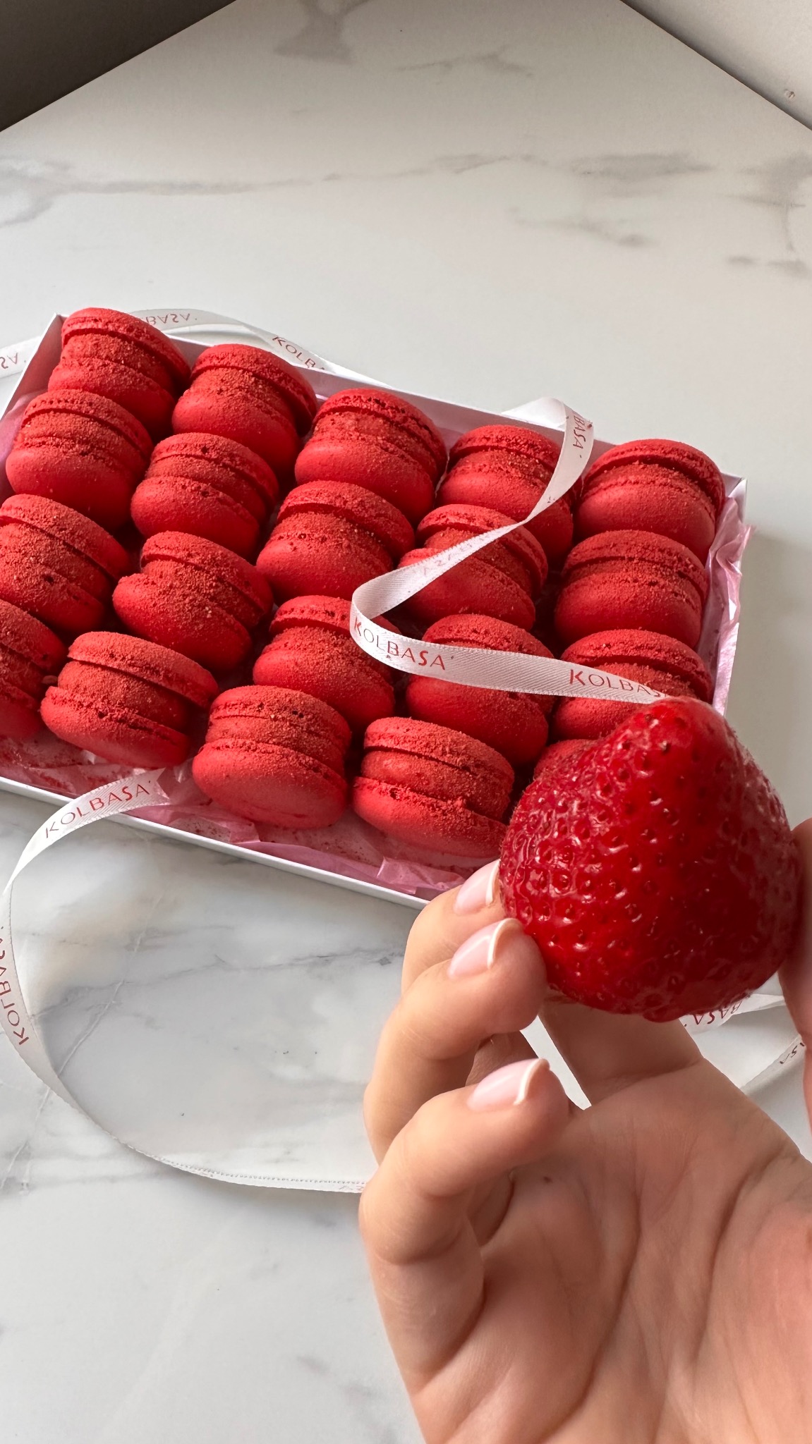 Pudełko czerwonych makaroników z wstążką z napisem i truskawką trzymaną w dłoni na marmurowym tle. Słodki upominek od cukierni.