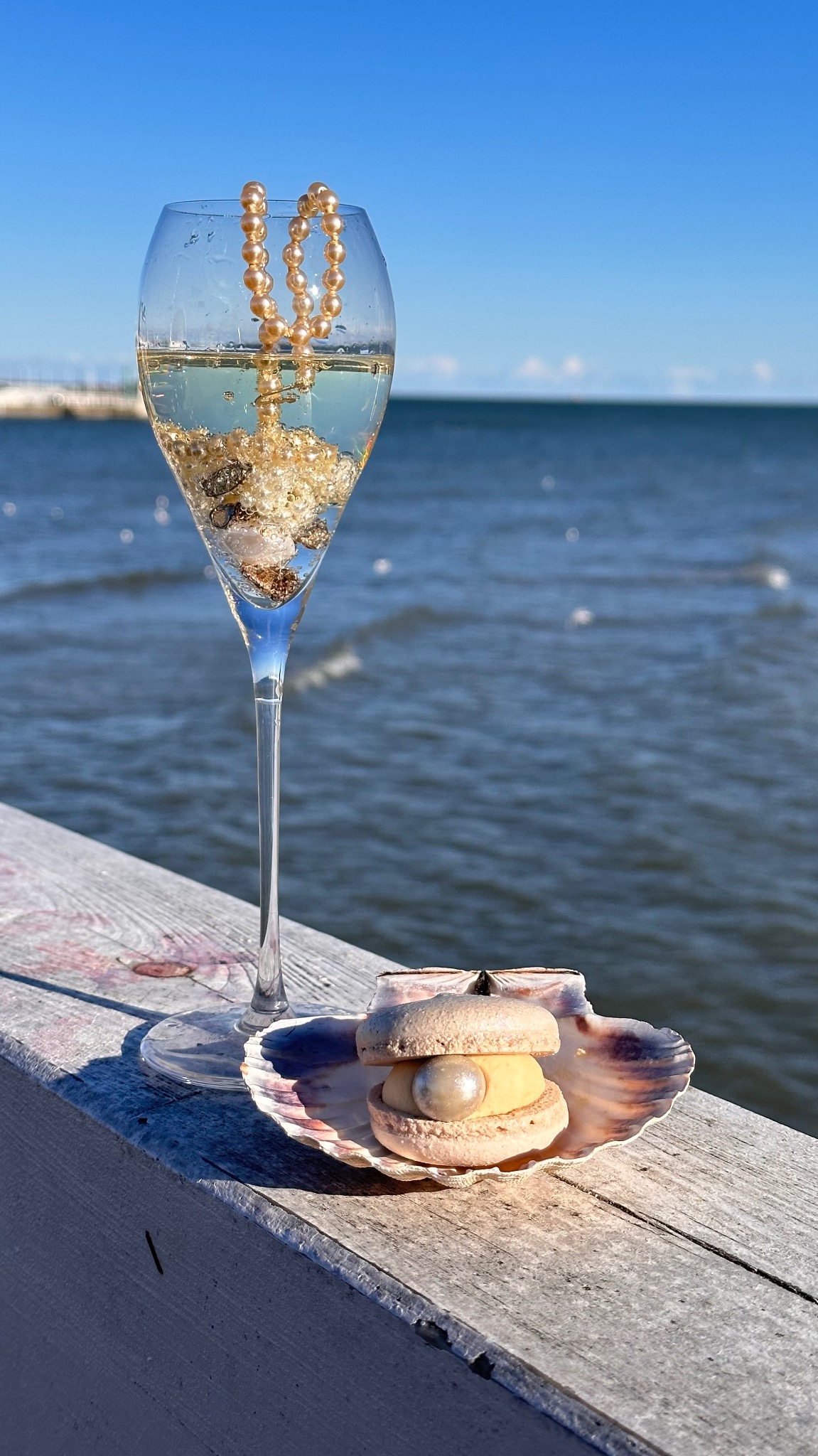 Dekoracyjny makaronik z perłą w muszli i kieliszek z perłami na tle morza, w słoneczny dzień. Nietypowa kompozycja cukiernicza z akcentem morskim.