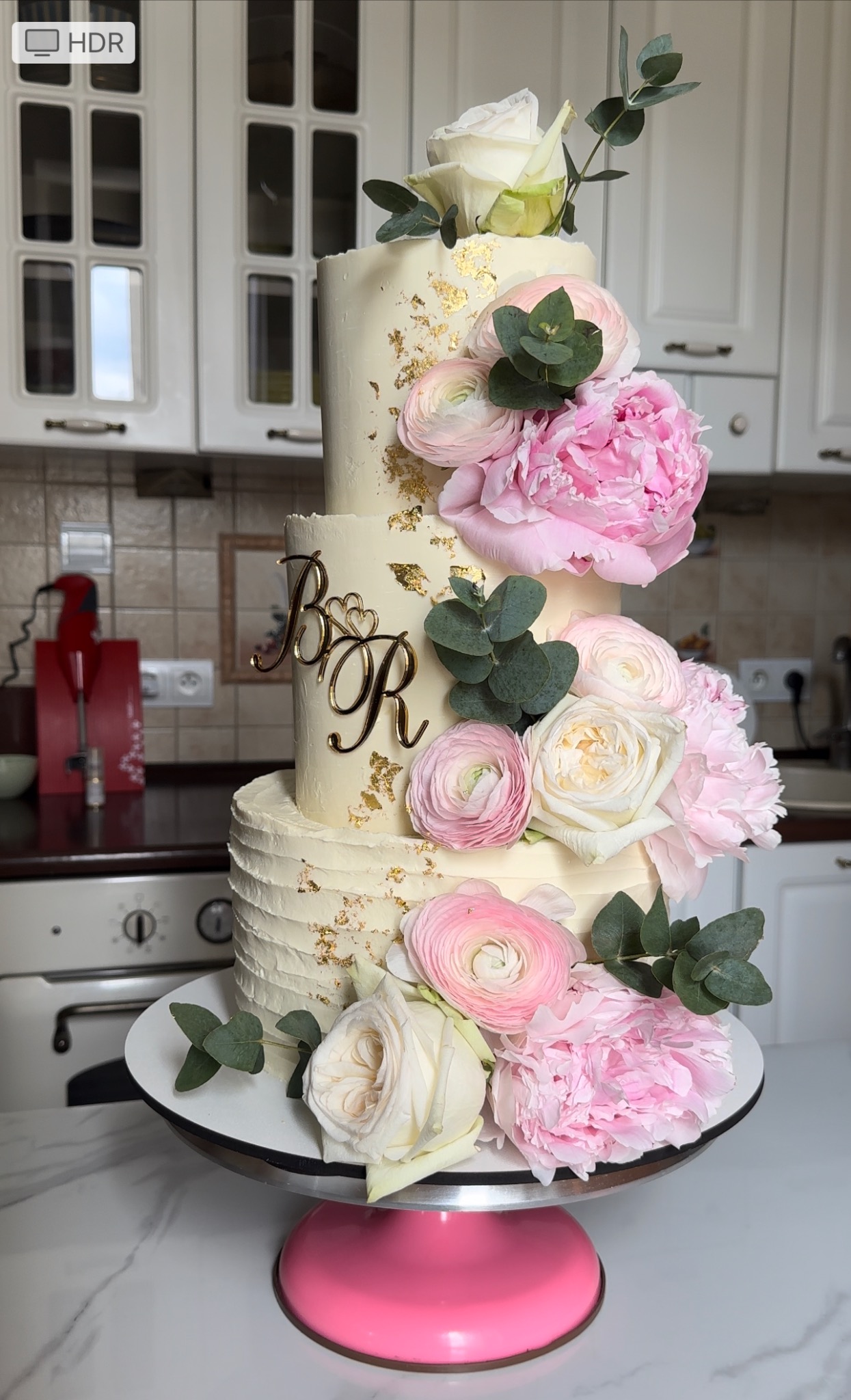 Elegancki, piętrowy tort weselny, zdobiony żywymi różowymi i białymi kwiatami, złotymi płatkami i inicjałami na tle białej kuchni.