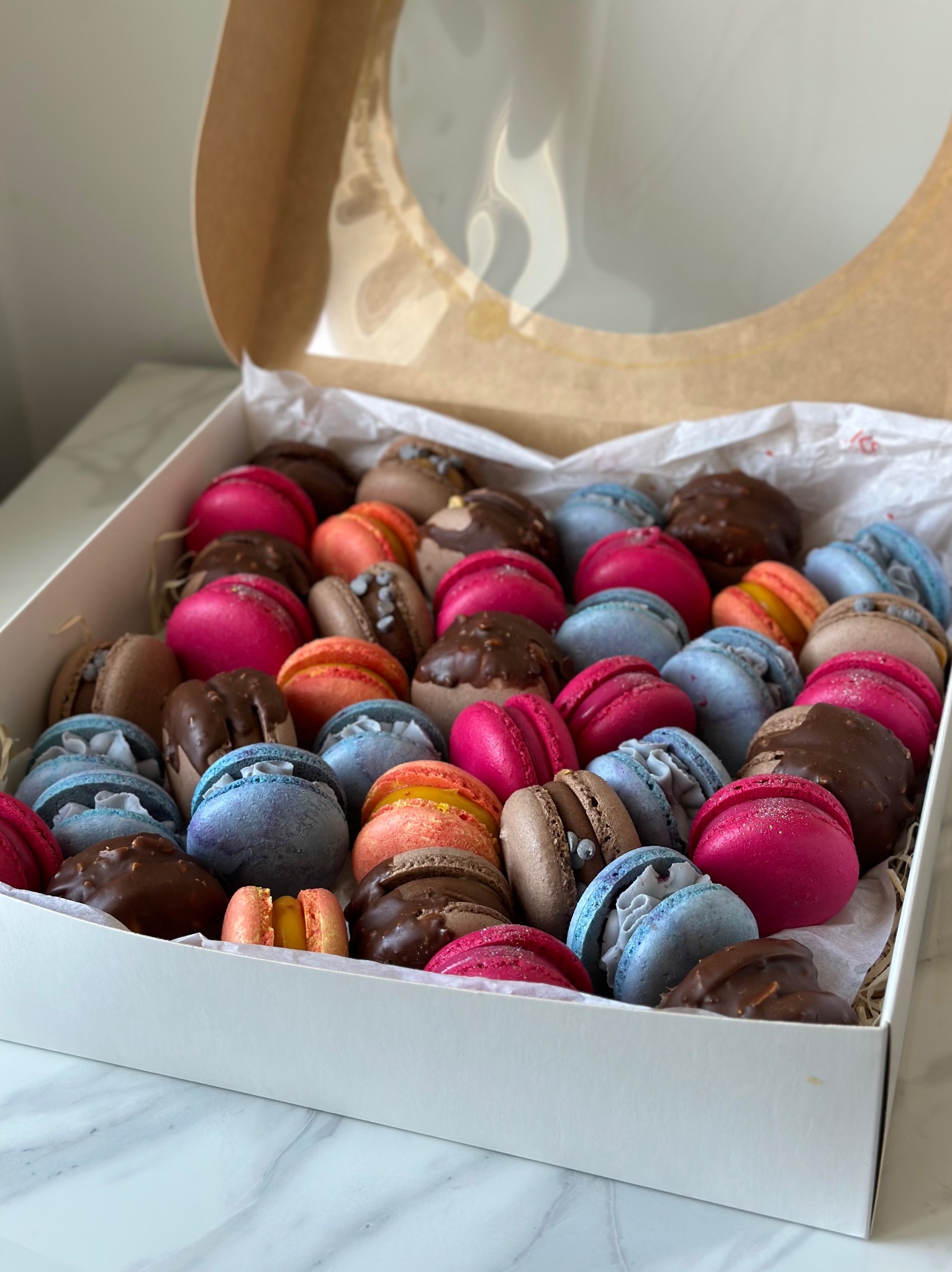 Kolorowe makaroniki w pudełku z okienkiem, różne smaki i polewy, na białym tle. Widok z góry, pudełko otwarte.