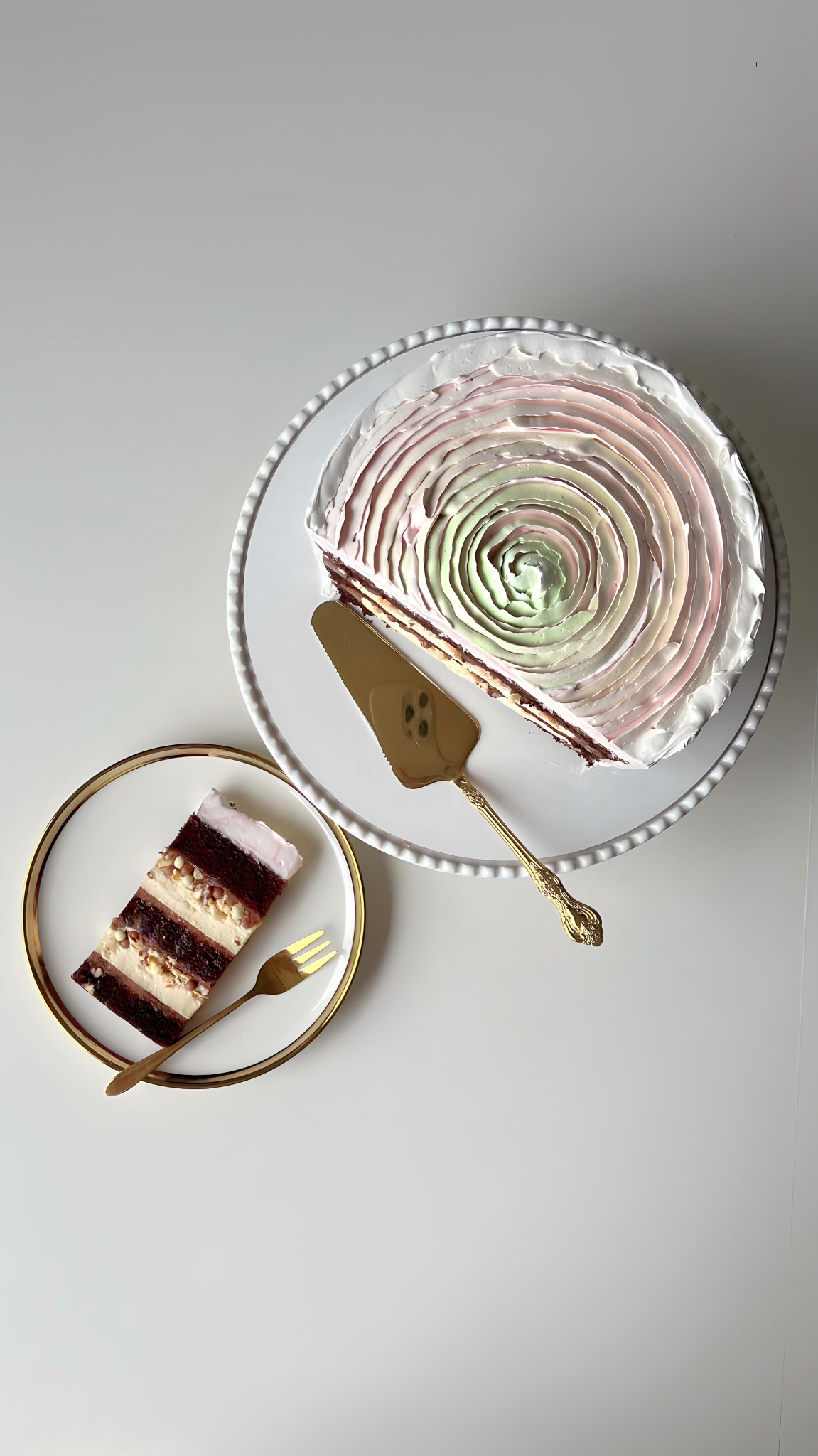 Kawałek tortu z kremem w pastelowych kolorach na talerzyku z widelcem obok tortu na paterze ze złotą łopatką. Ujęcie z góry na białym tle.