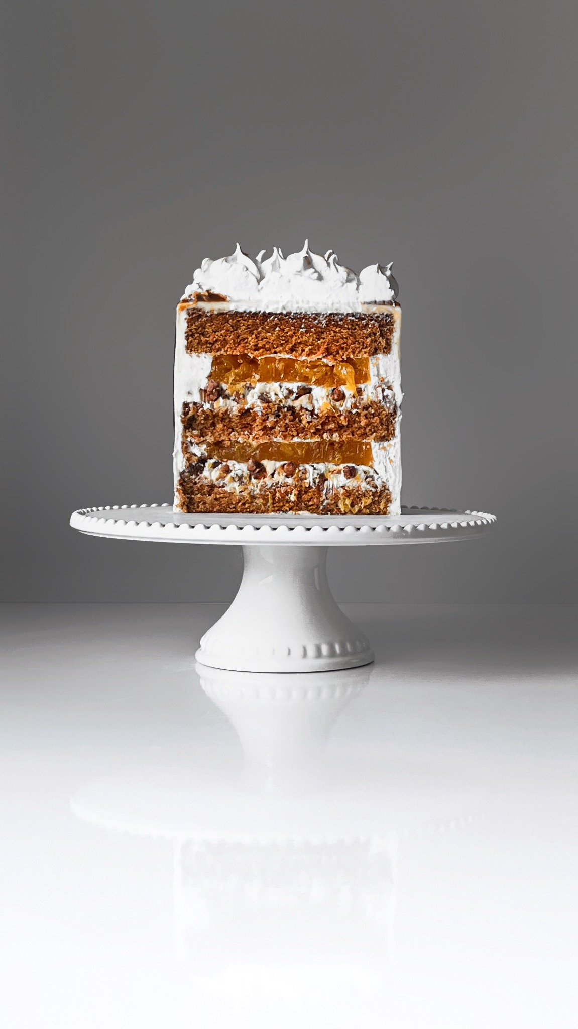 Przekrój tortu na białym, eleganckim stojaku. Widoczne warstwy ciasta, kremu i owocowej galaretki, całość zwieńczona bitą śmietaną. Minimalistyczne tło.