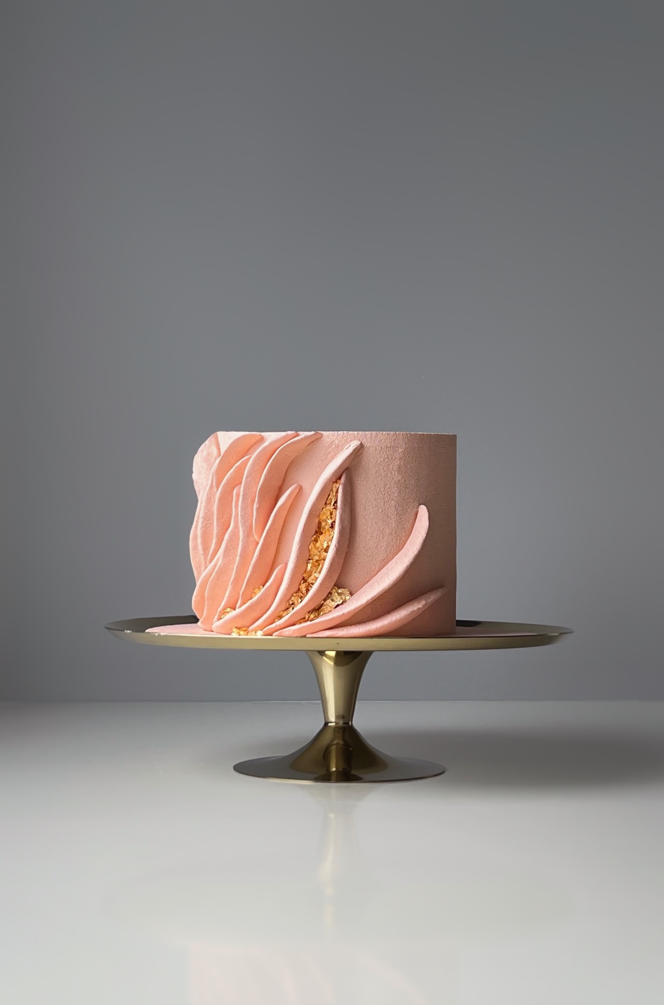 Delikatny tort w odcieniach różu z abstrakcyjną dekoracją z płatków złota, umieszczony na złotej paterze. Minimalistyczne tło podkreśla elegancję.