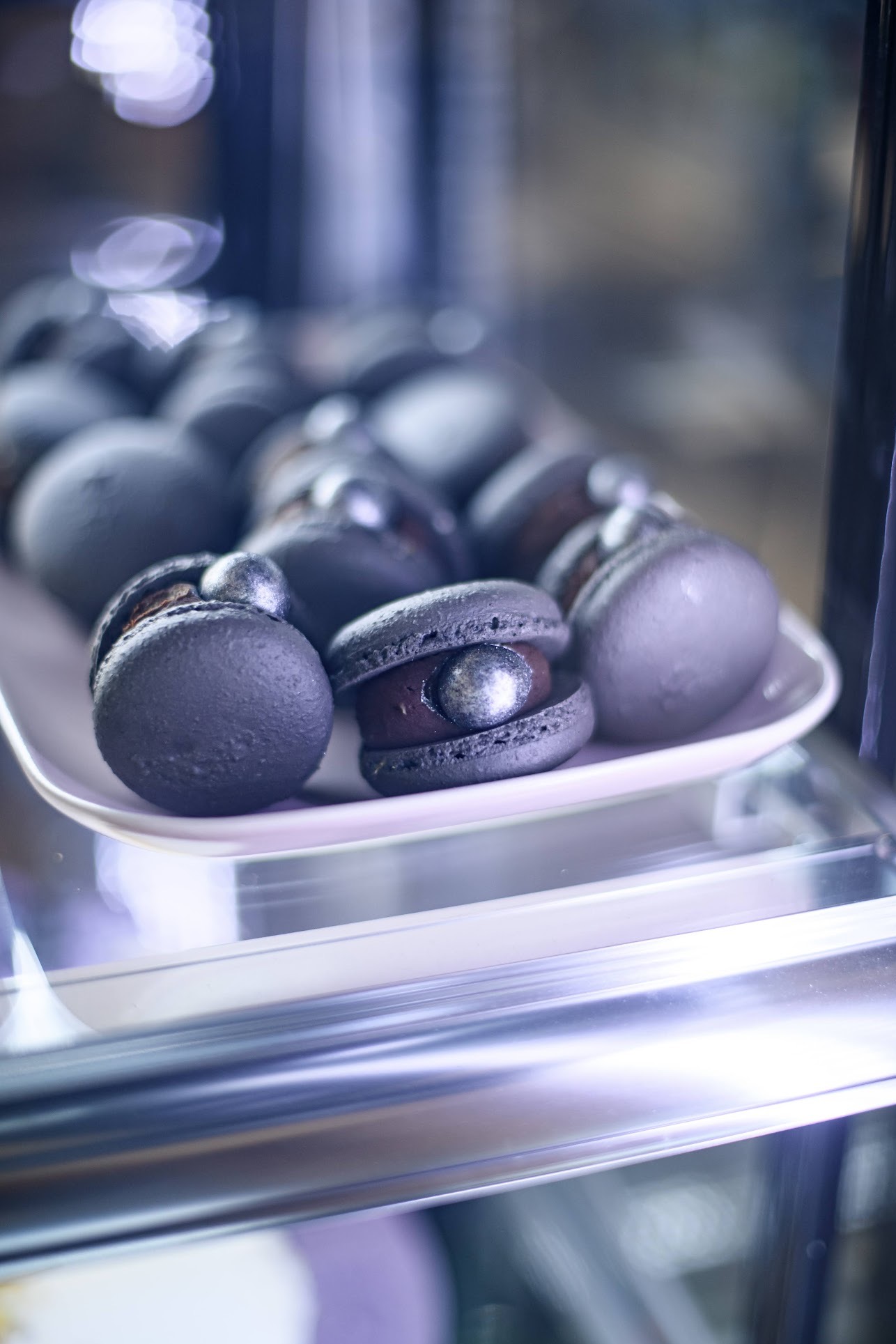 Ciemne, artystyczne makaroniki z błyszczącymi srebrnymi akcentami na białym talerzu w chłodnej witrynie, prezentowane w cukierni.