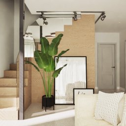 ARCHITEKT MAŁGORZATA RUSINIAK MR ATELIER - Jasne wnętrze z drewnianymi schodami, rośliną w czarnej donicy i minimalistycznymi plakatami. Widoczne oświetlenie punktowe i fragment sofy z poduszkami.