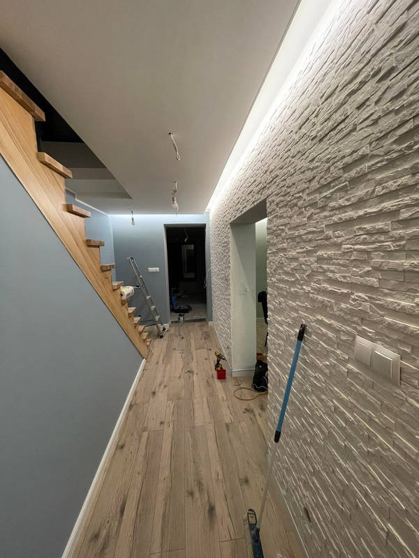 Korytarz po remoncie: drewniane schody, panelowa podłoga, ściana z białej cegły dekoracyjnej z oświetleniem LED. Widoczna drabina i narzędzia.