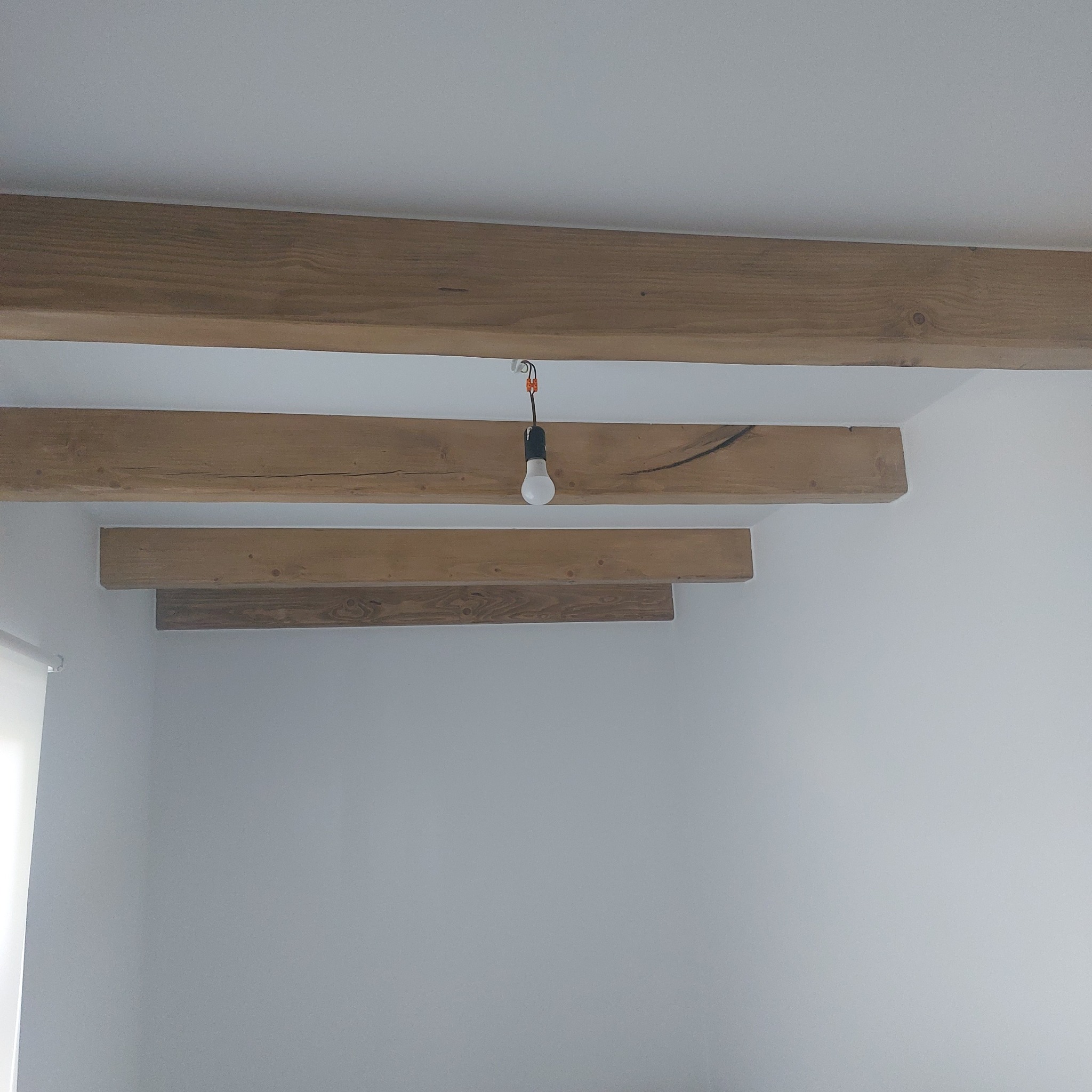 Drewniane belki stropowe w jasnym wnętrzu, widoczne ślady po sękach i naturalne przebarwienia drewna. Wisząca żarówka na kablu dodaje surowego charakteru.