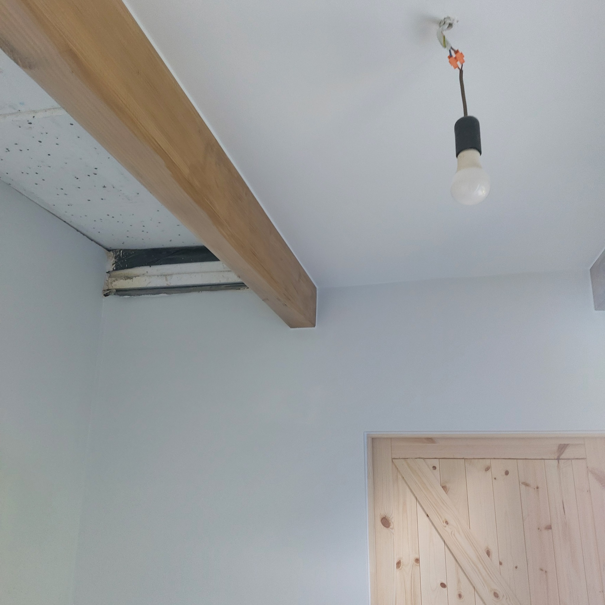 Jasne wnętrze w trakcie wykańczania: drewniana belka pod sufitem, surowa drewniana stolarka drzwiowa, ściany pomalowane na jasny kolor, wisząca żarówka.