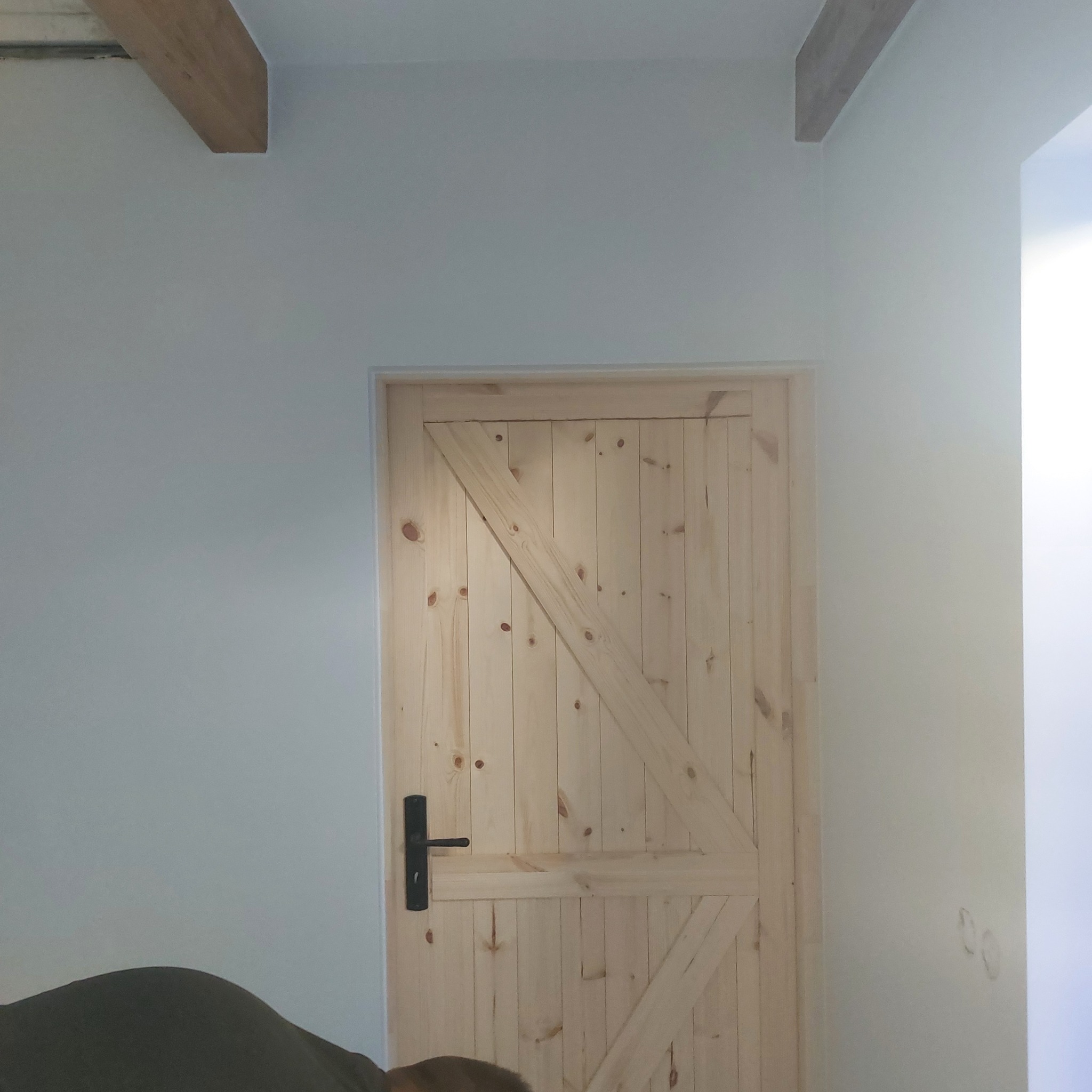 Jasne drewniane drzwi w stylu rustykalnym z czarną klamką na tle pomalowanej ściany i drewnianych belek stropowych. Wykończenie wnętrza.