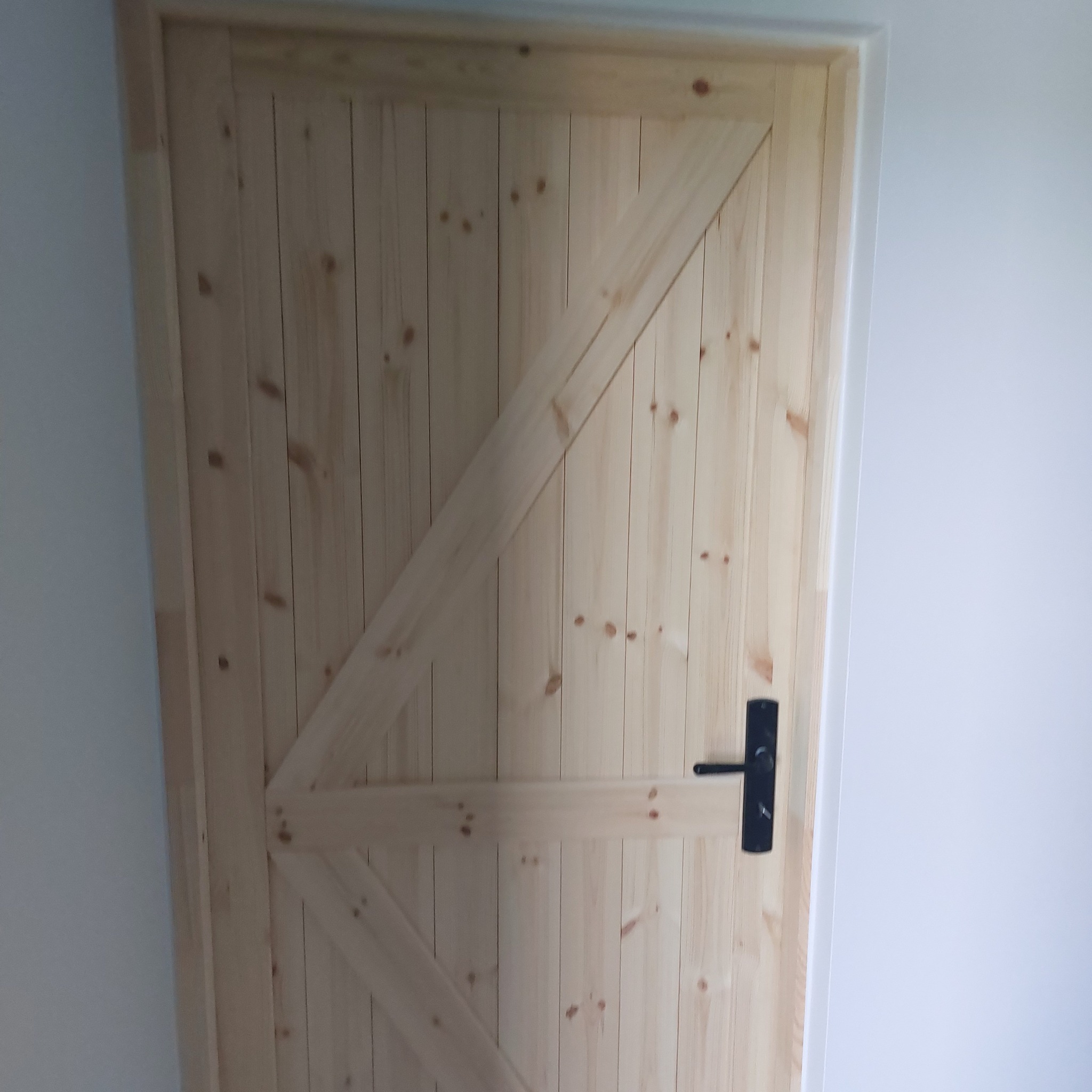 Jasne, drewniane drzwi wewnętrzne z czarną klamką, widoczne na tle białej ściany. Charakterystyczny wzór desek w jodełkę. Prosty, minimalistyczny design.