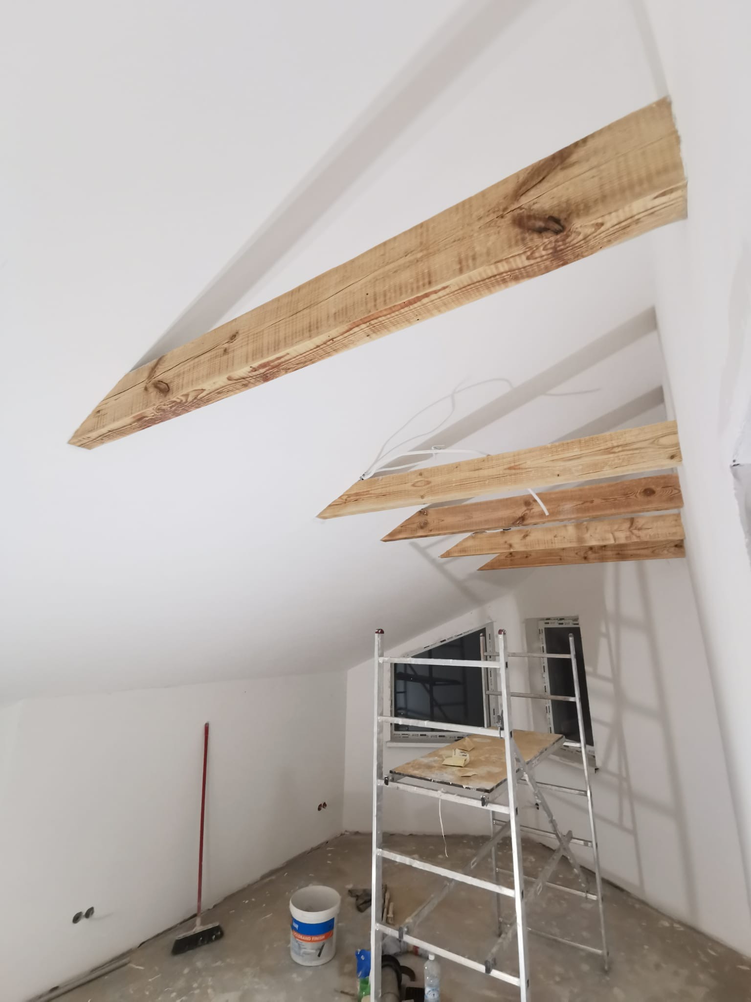 Poddasze w trakcie prac wykończeniowych: drewniane belki dekoracyjne pod sufitem, rusztowanie, ściany przygotowane do malowania. Widoczne okna i akcesoria malarskie.