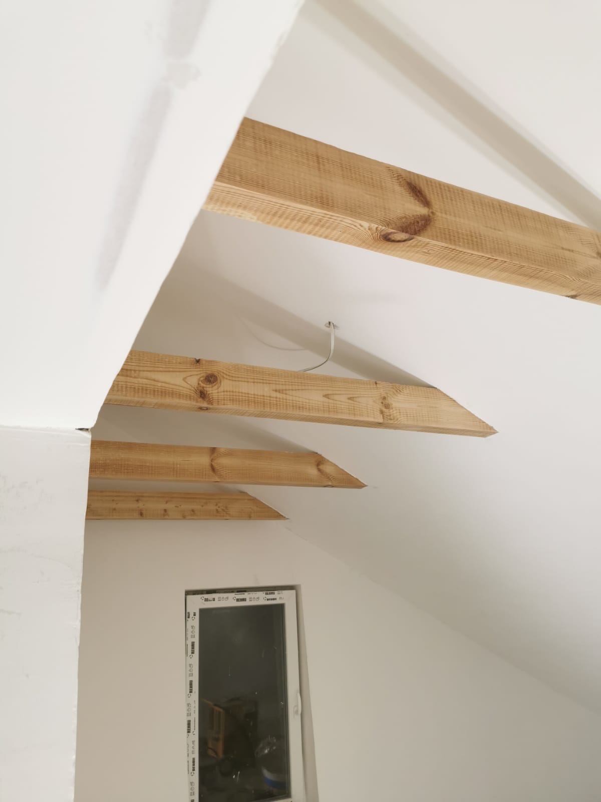 Białe poddasze z drewnianymi belkami stropowymi, widoczne okno. Belki zamontowane w równych odstępach. Sufit i ściany świeżo pomalowane na biało.