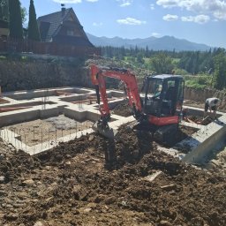 Iwona Knapczyk - Fundamenty domu z małą koparką Kubota w akcji, malownicze góry w tle. Widoczne zbrojenia i szalunki. Prace ziemne w toku, słoneczny dzień.
